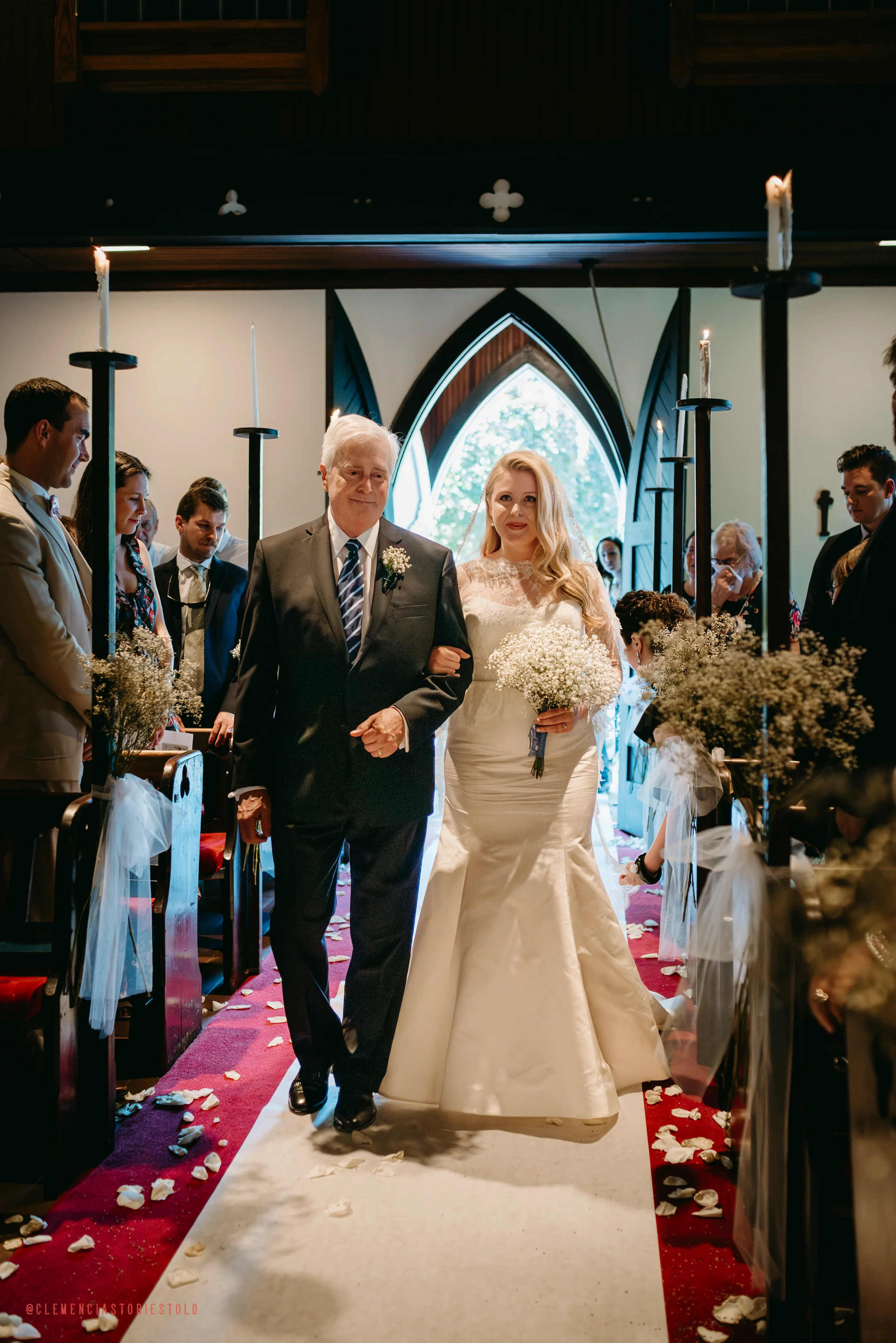 A bride walking down the aisle with her father during a wedding ceremony inside a church, with guests sitting on either side and wedding decorations visible.