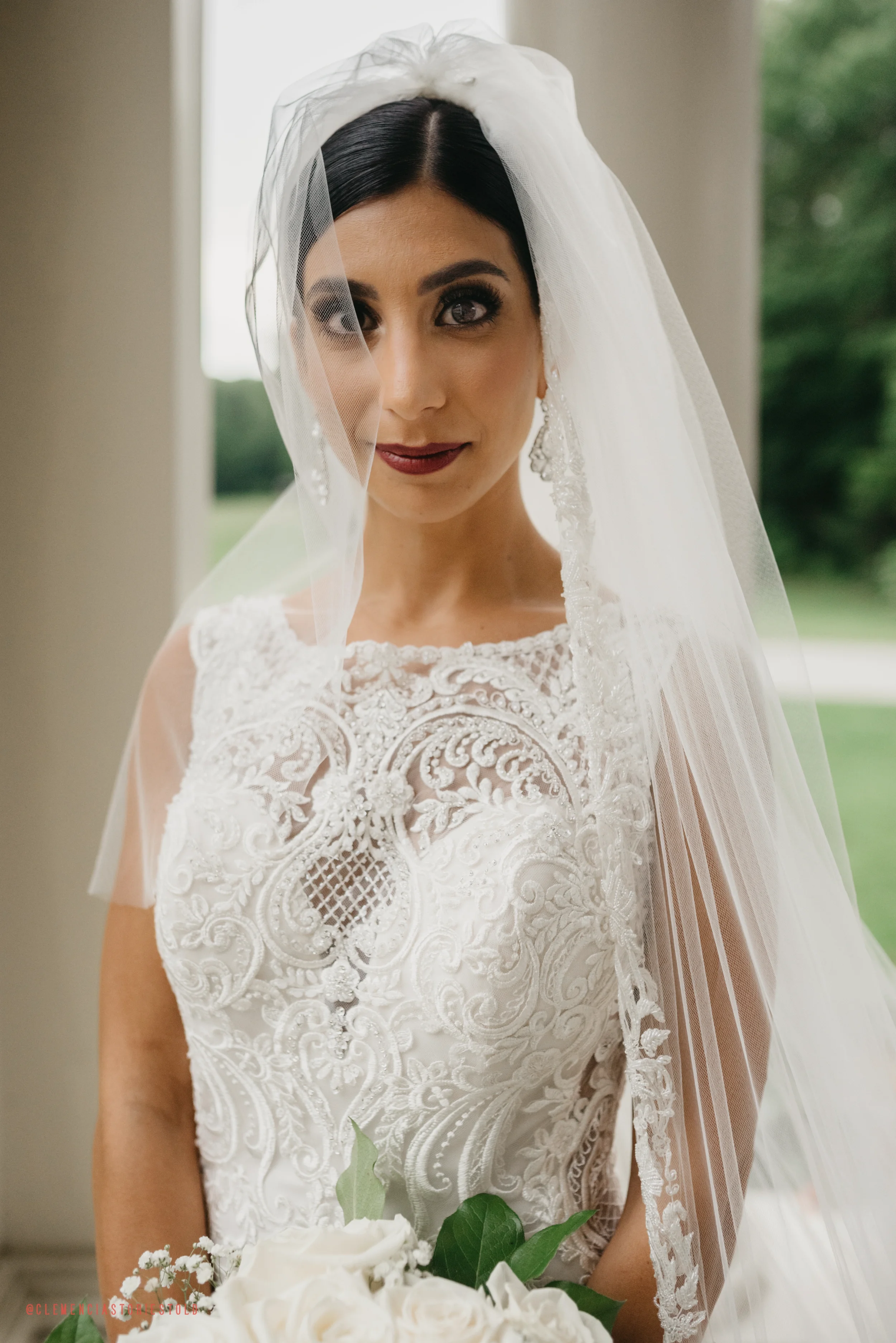 A bride with dark hair wears a lace wedding gown and veil, holding a bouquet of white roses, standing indoors with greenery visible outside.