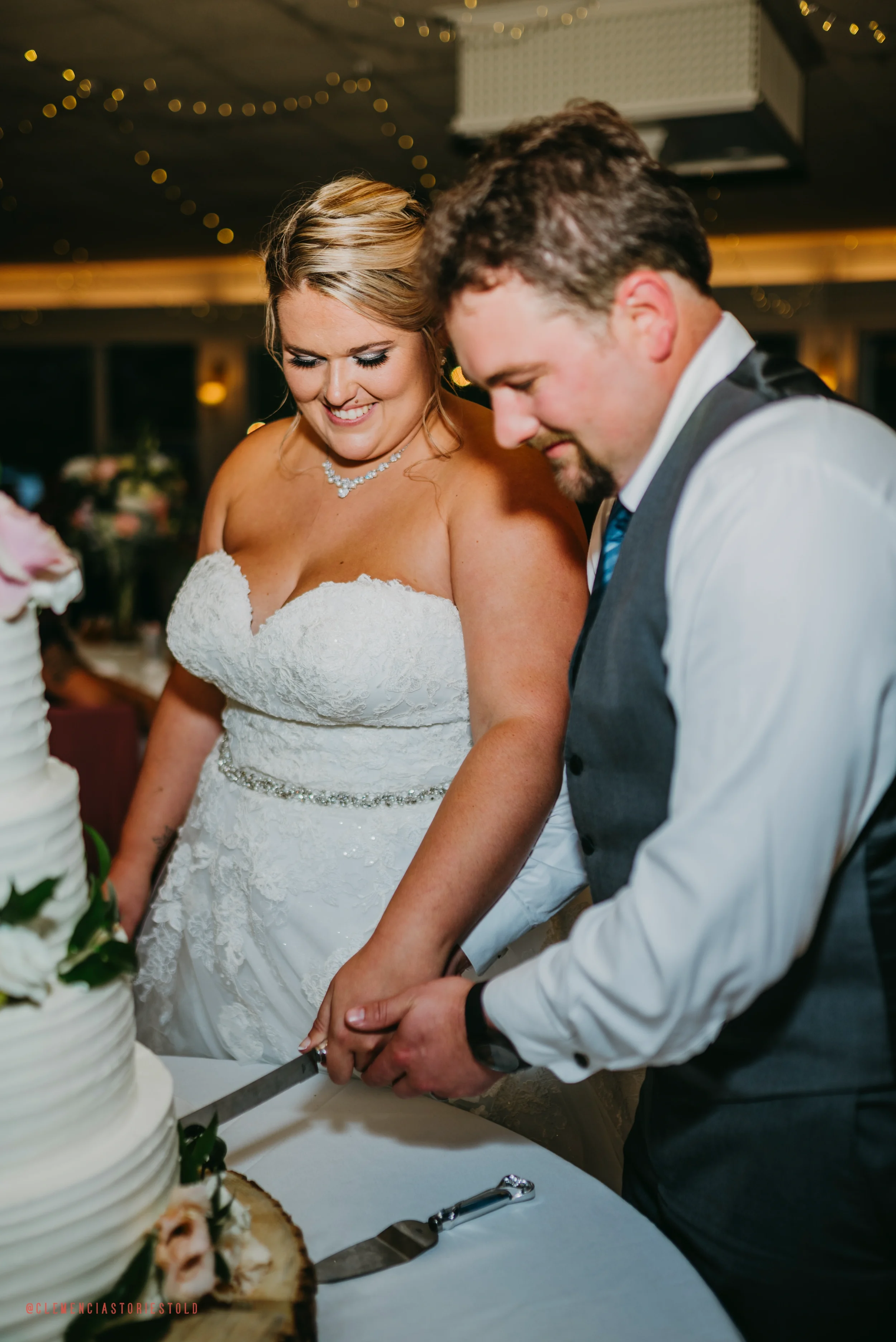 A bride and groom share a moment while cutting their wedding cake together, smiling and holding the knife with both hands, in a decorated reception hall.