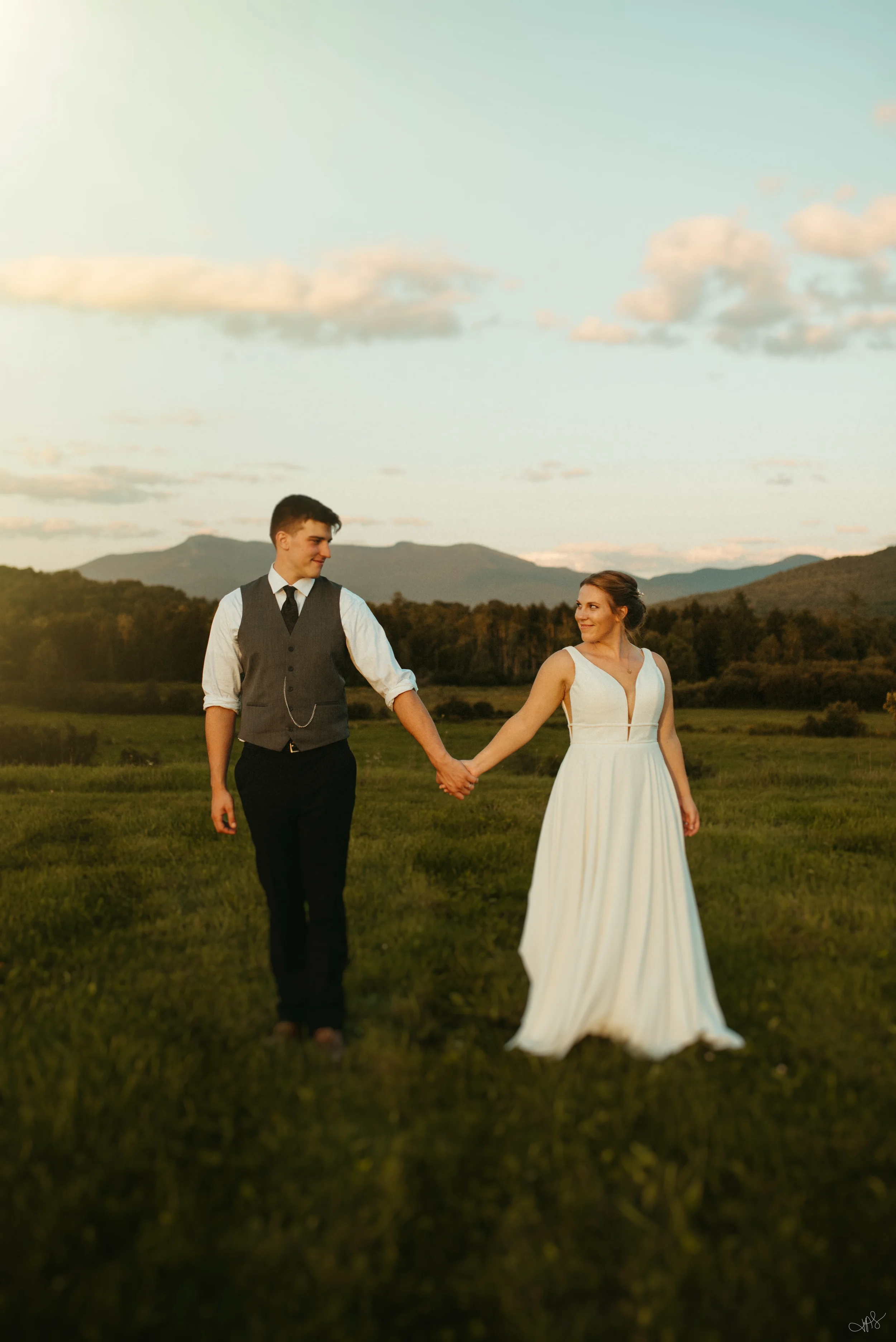 A young couple holding hands while walking in a grassy field during sunset, with mountains and a partly cloudy sky in the background.