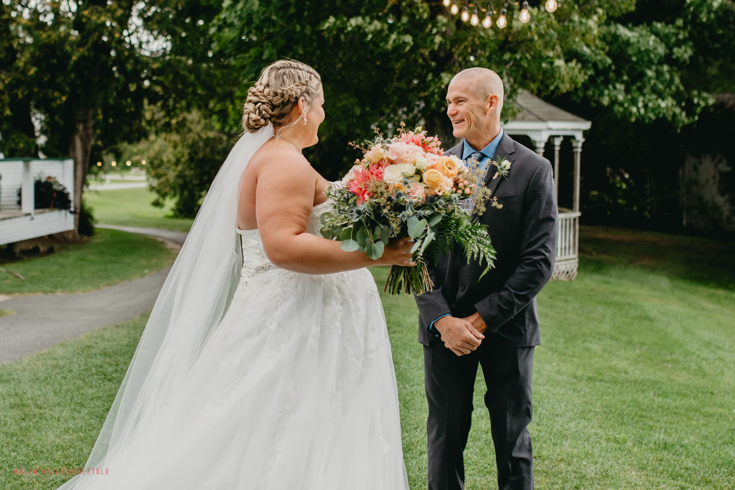Bride in a white wedding dress holding a bouquet of flowers, smiling at a man in a black suit, outdoors with trees and a gazebo in the background.