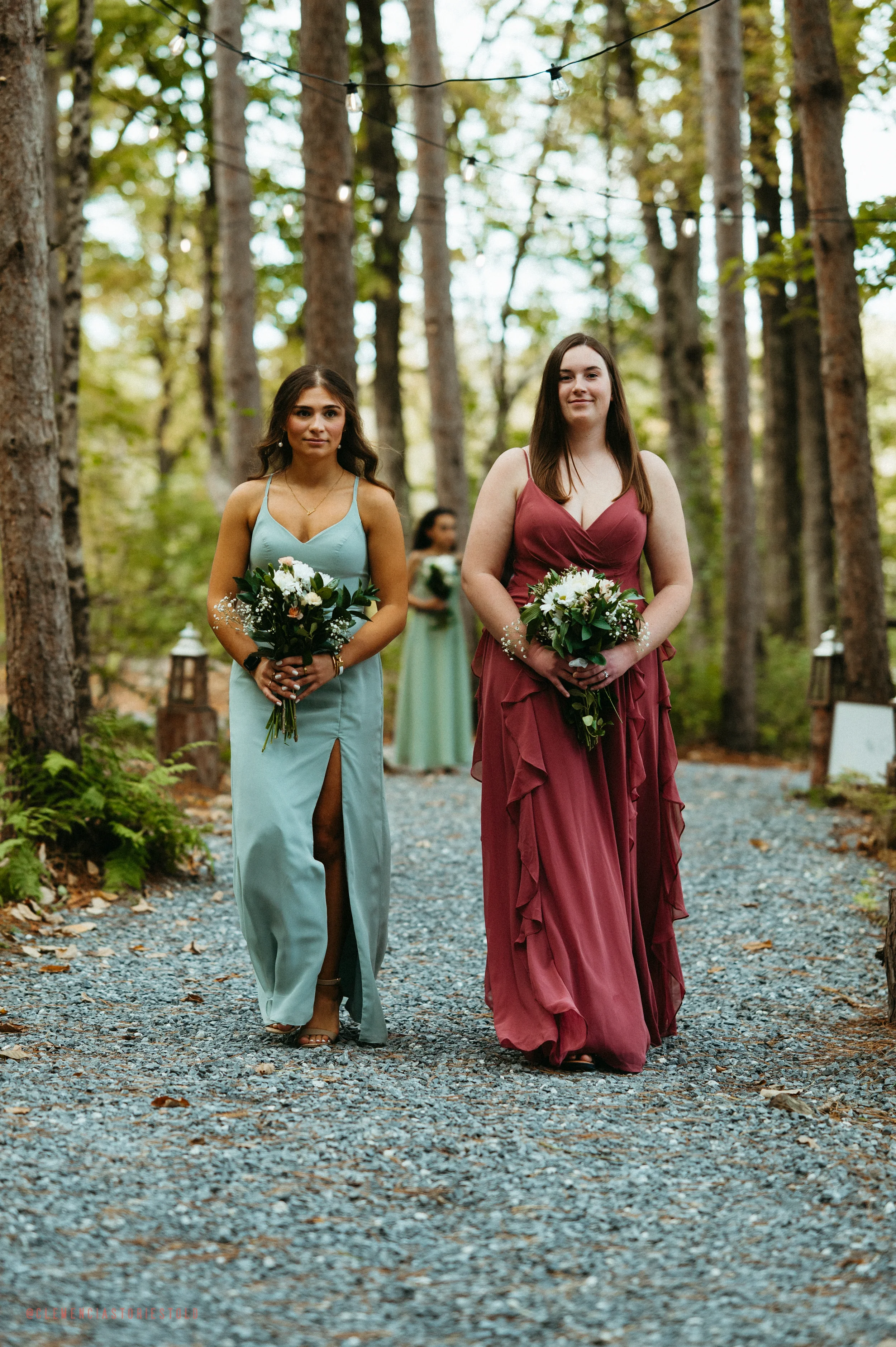 Two women in formal dresses holding bouquets walking down a forest path decorated with string lights, with a third woman in the background.