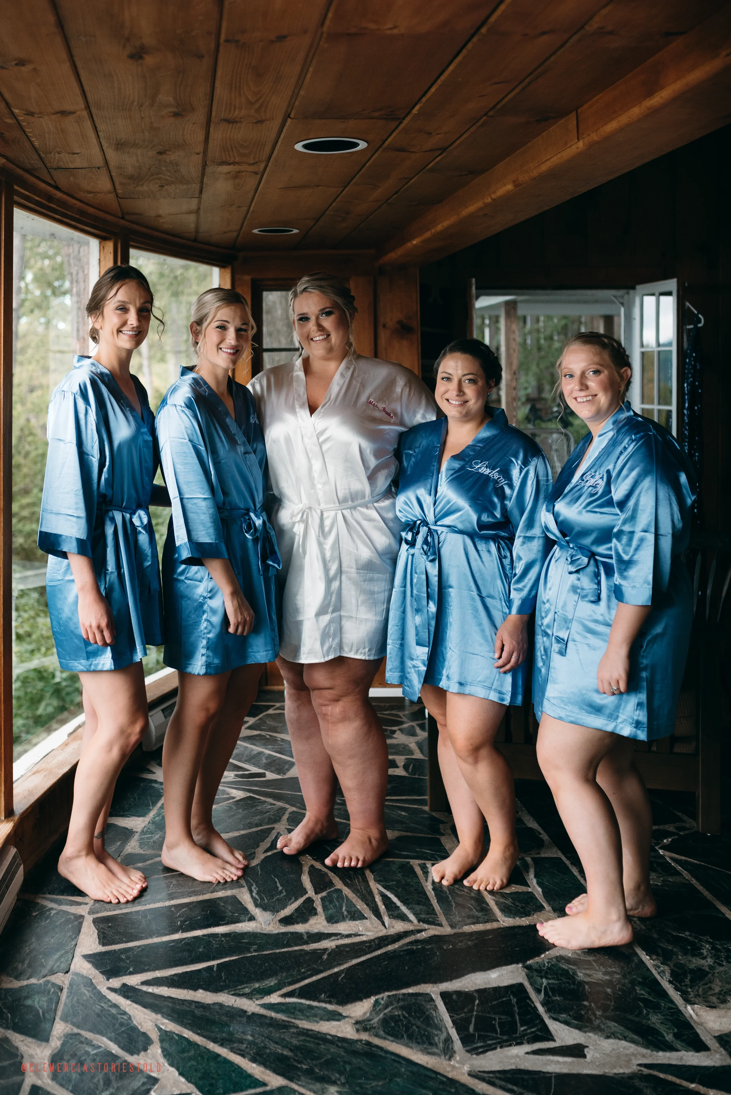 Five women in satin robes, four in blue and one in white, standing barefoot on a patterned stone floor inside a wooden room with large windows. They are smiling and posing for a photo, likely prior to a wedding.