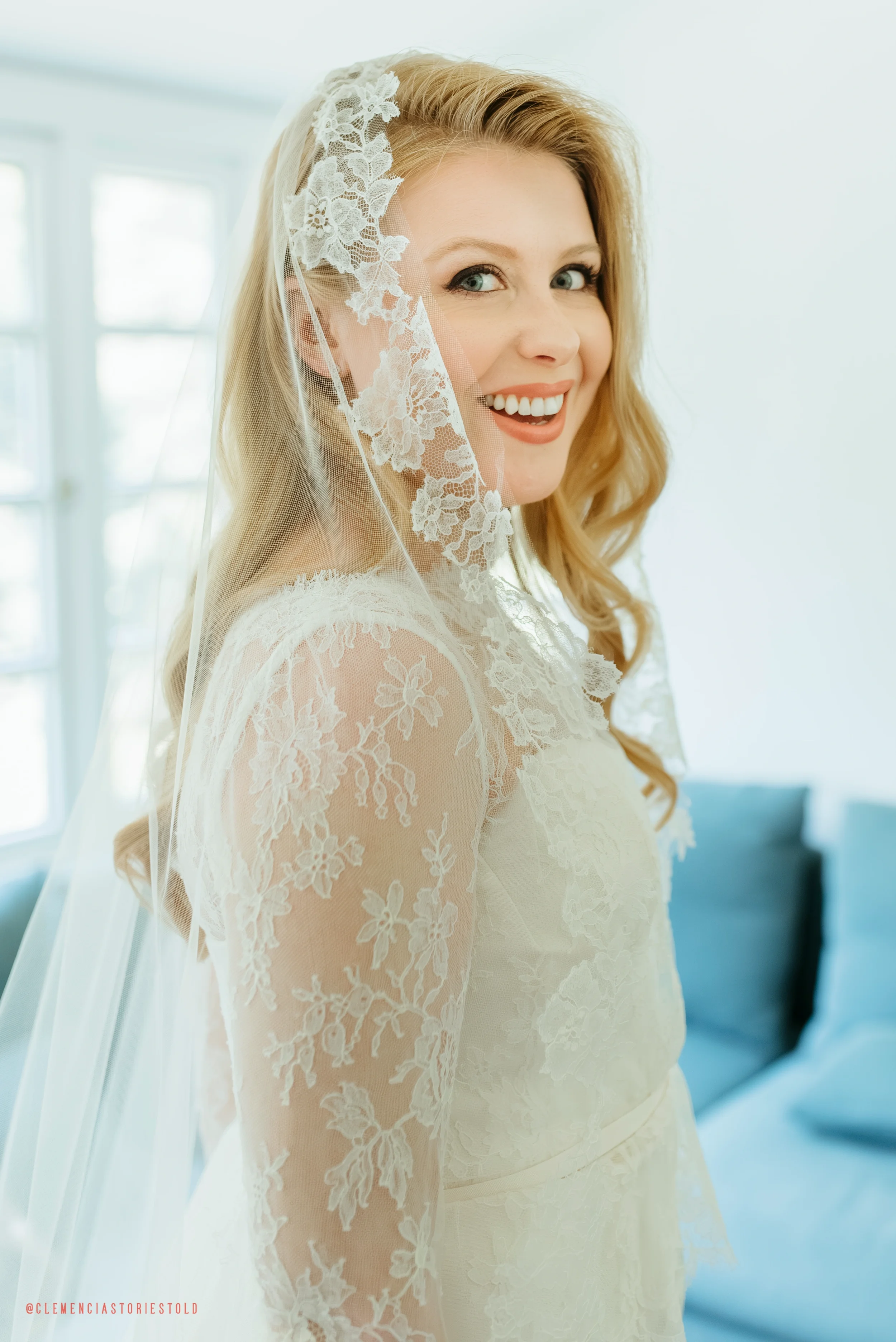 A woman smiling, wearing a lace wedding dress and a lace veil, standing indoors near a window.