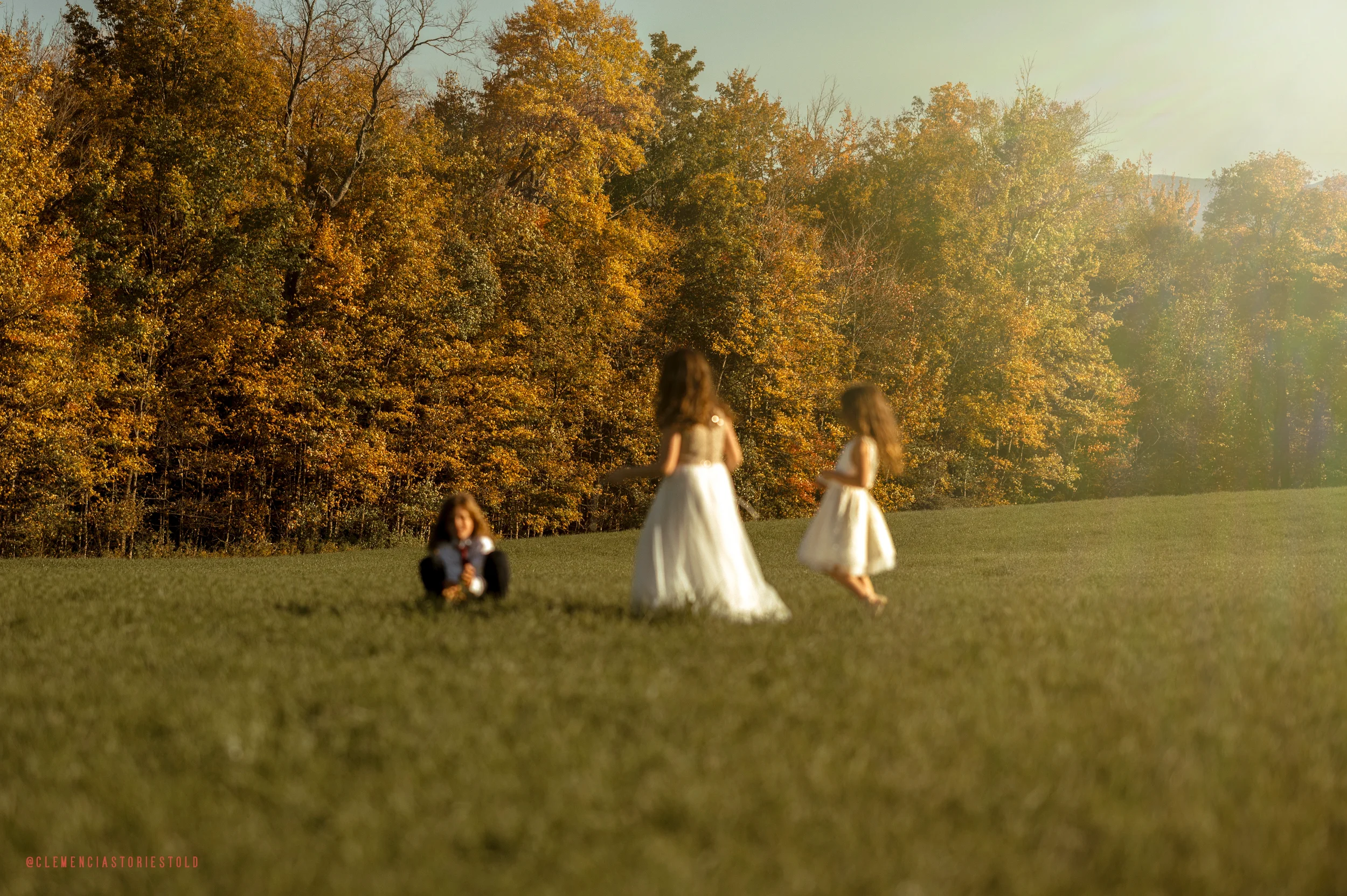 Three girls in white dresses playing on a grassy field with an autumn forest background, during golden hour sunlight.