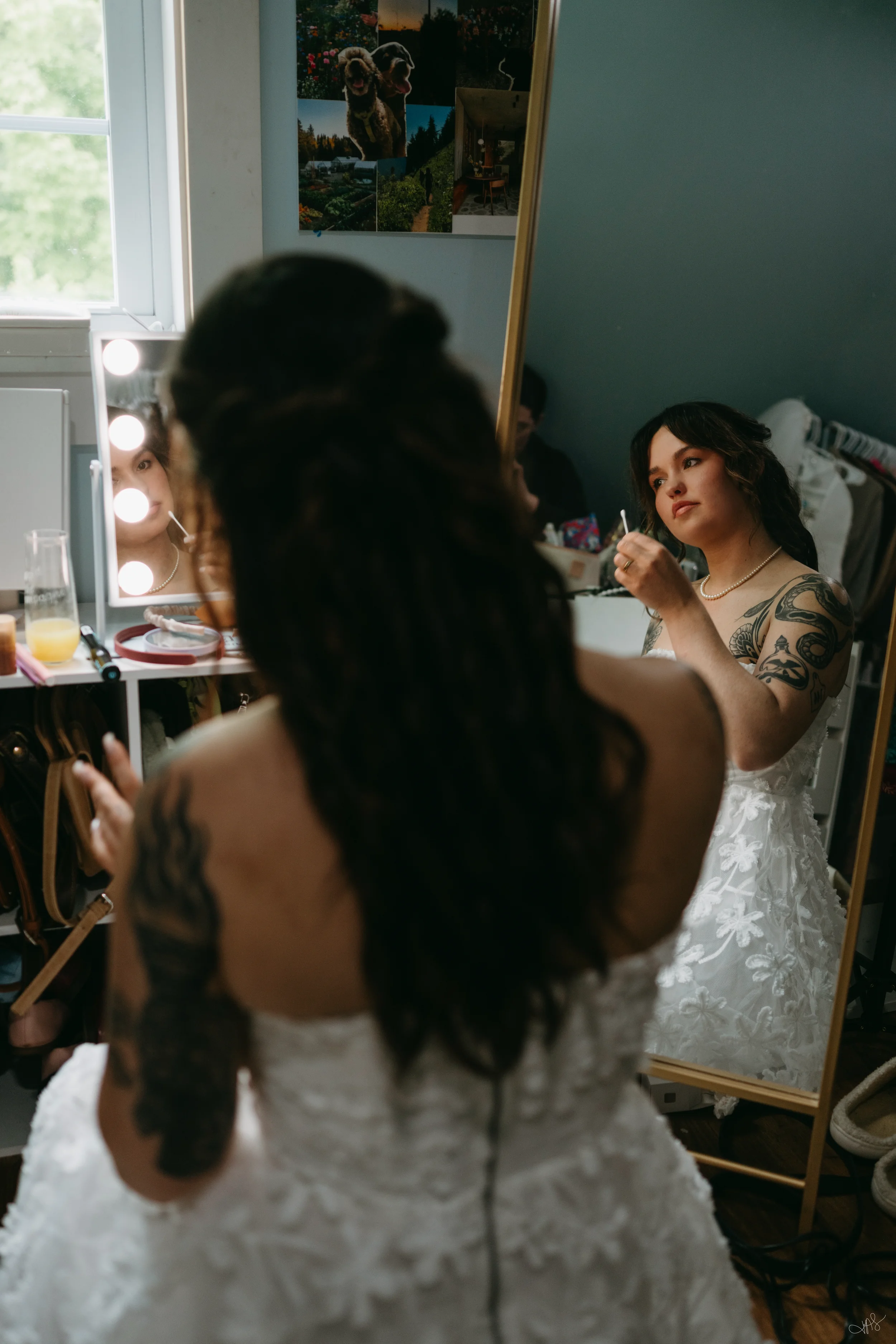 A woman dressed in a white wedding gown is sitting in front of a mirror, getting ready. She has tattoos on her shoulders and dark wavy hair. In the foreground, another woman with long dark hair, also with tattoos, is seated with her back to the camera, possibly doing her makeup or hair. The room is decorated with a photo collage of dogs, nature, and a house on the wall. There is a window to the left with natural light coming in.