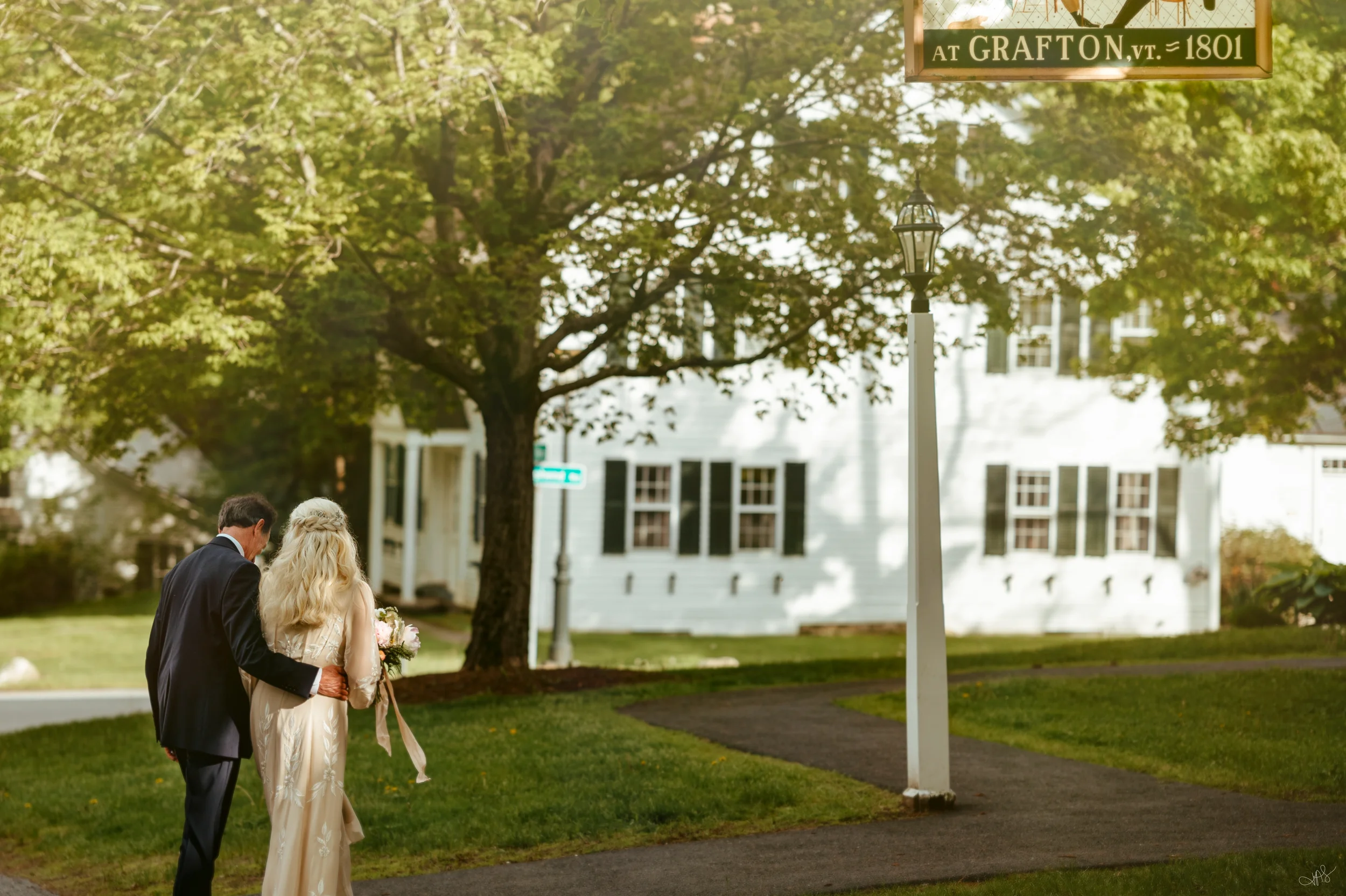 A man and woman dressed in wedding attire walking outdoors on a lush green lawn, with trees and white houses in the background. There is a street sign that reads 'at Grafton, VT, 1801' and a lamp post.