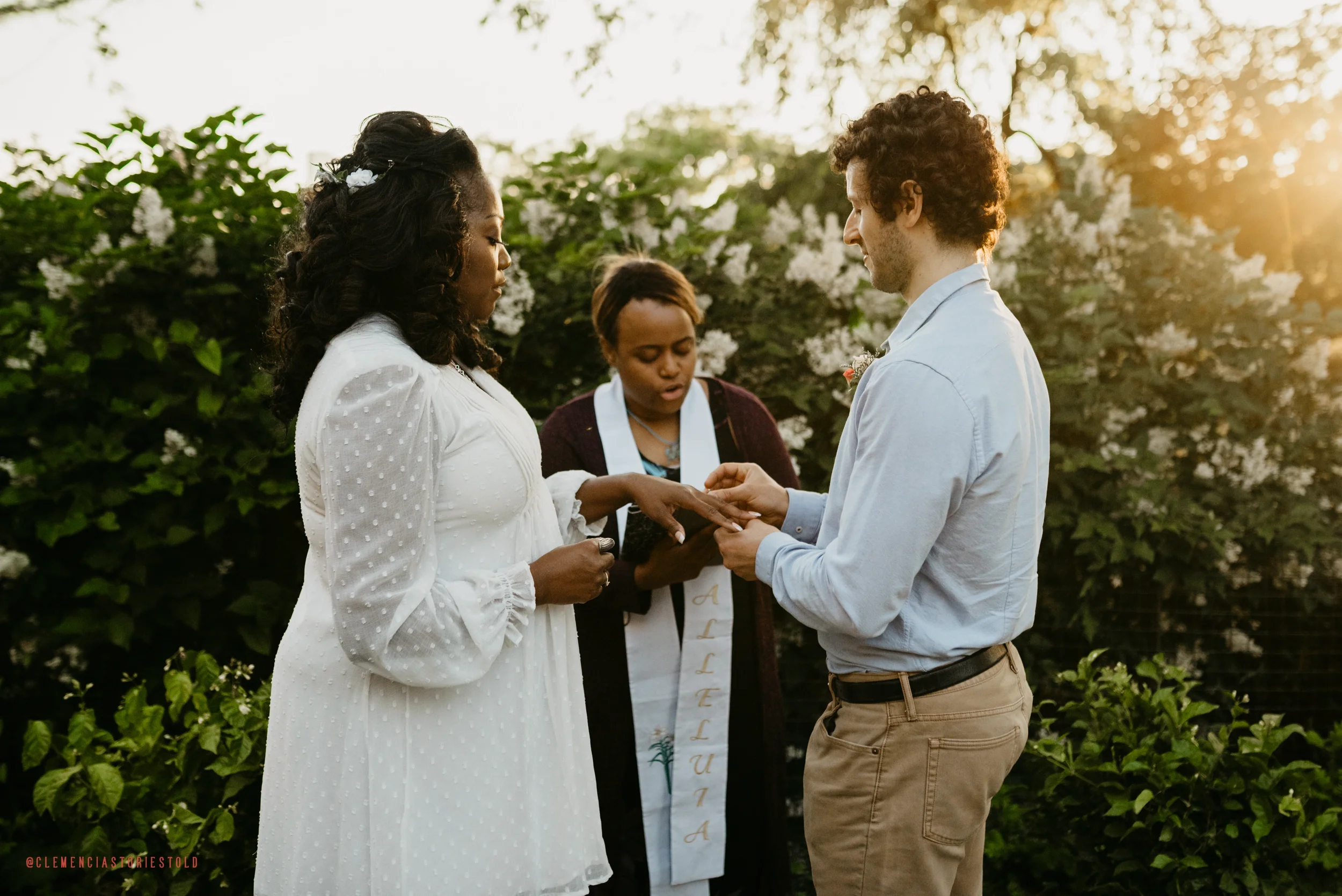 A couple exchanging wedding rings during an outdoor wedding ceremony at sunset, with an officiant standing behind them and lush greenery in the background.