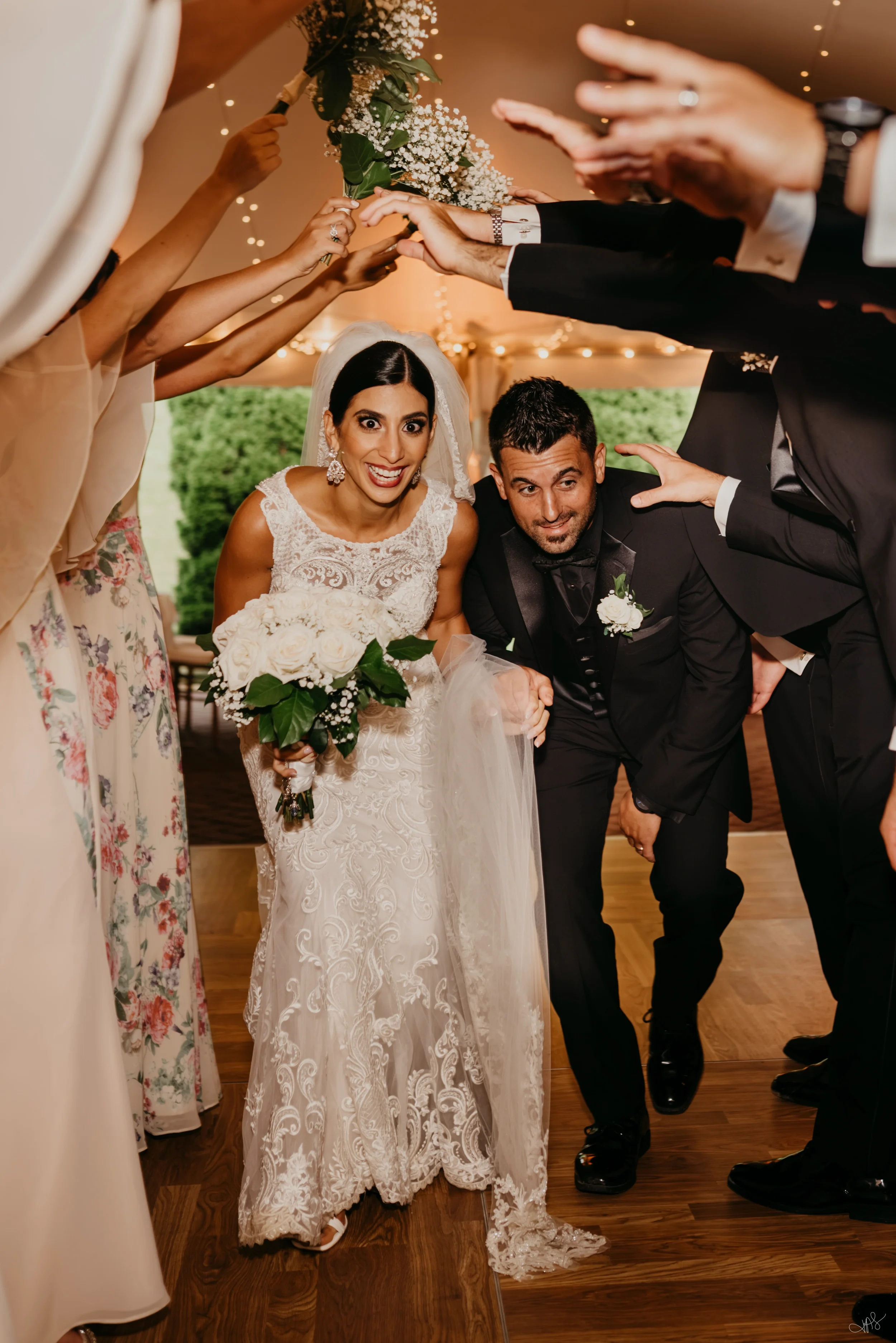 Bride and groom surrounded by friends and family forming a tunnel with hands and flowers during a wedding celebration