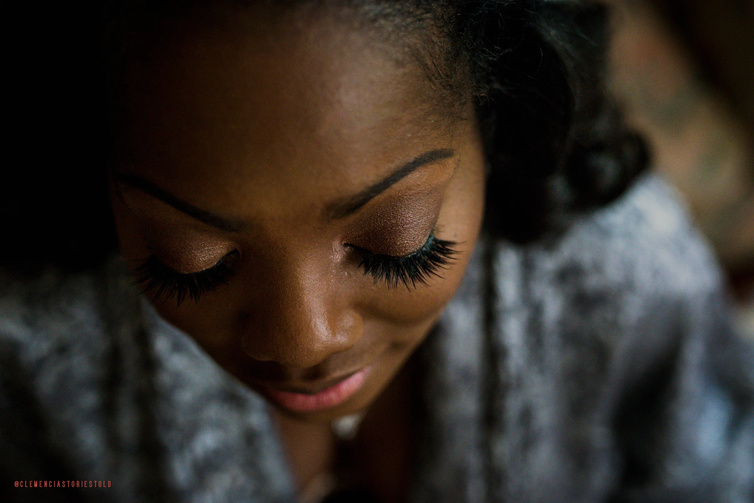 A close-up of a woman with dark hair and makeup, her eyes closed, showing long eyelashes and eyeshadow, wearing a gray top.