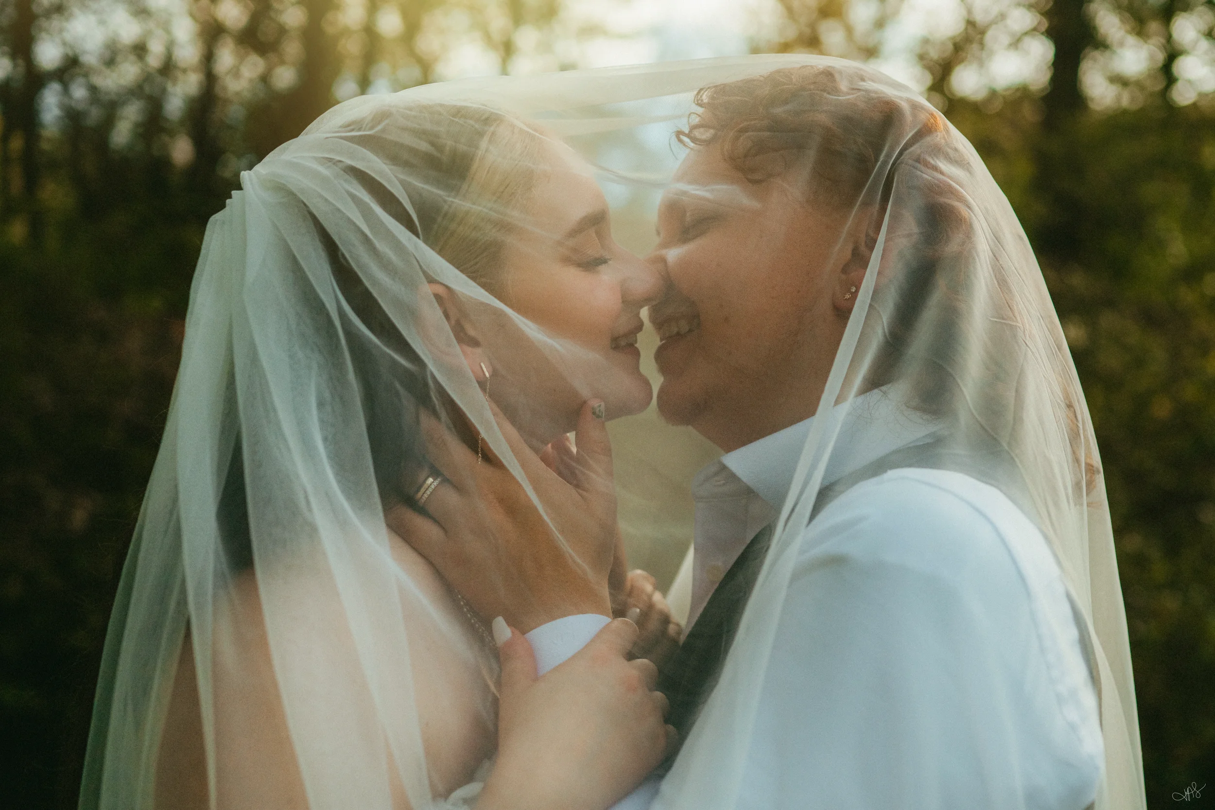 Two women, one with blonde hair and the other with curly hair, under a wedding veil sharing a kiss outdoors with sunlight filtering through trees.