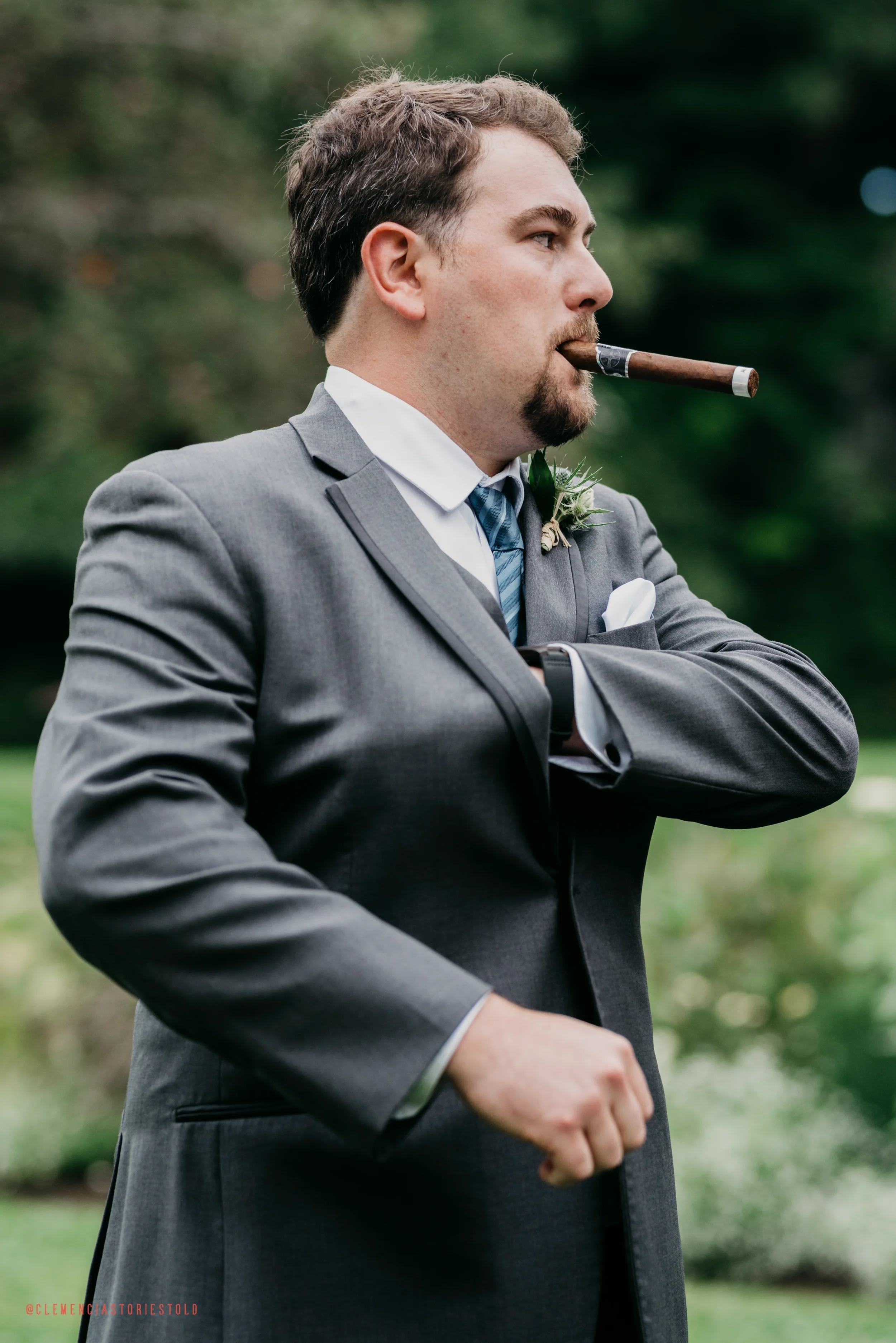 A man dressed in a gray suit with a boutonniere, wearing a blue striped tie, with a cigar in his mouth and a crossed-arm stance in an outdoor setting with blurred green trees in the background.