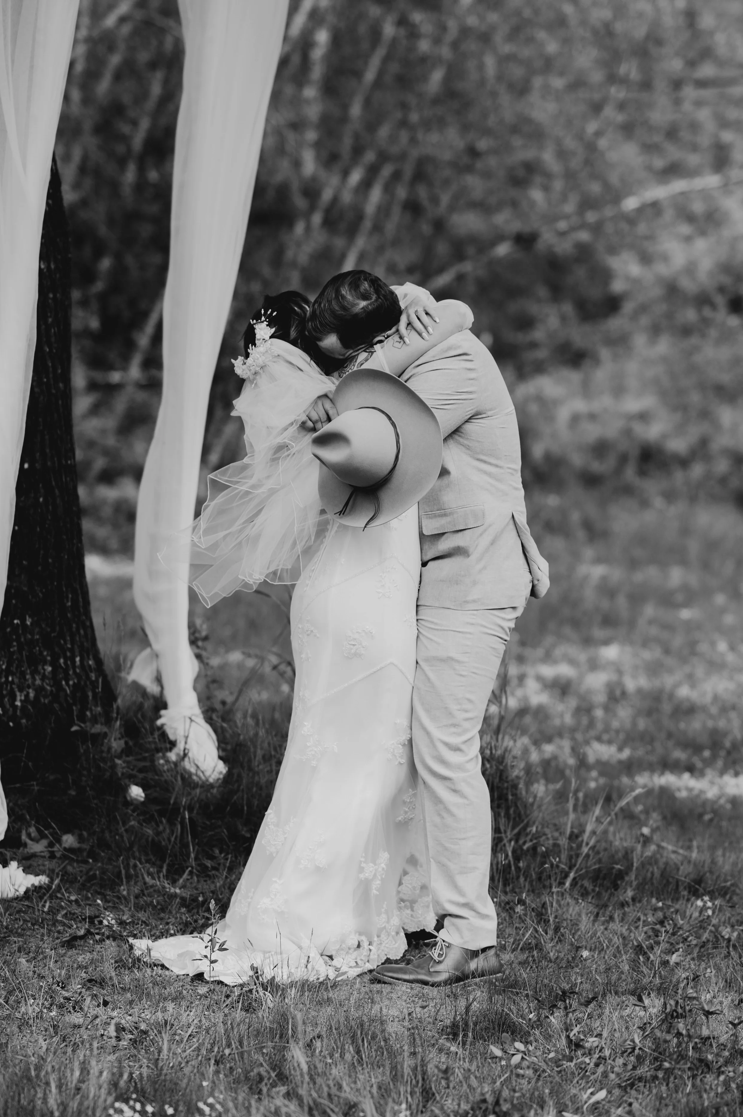 Black and white photo of a couple embracing outdoors during a wedding, with trees in the background and draped fabric nearby.