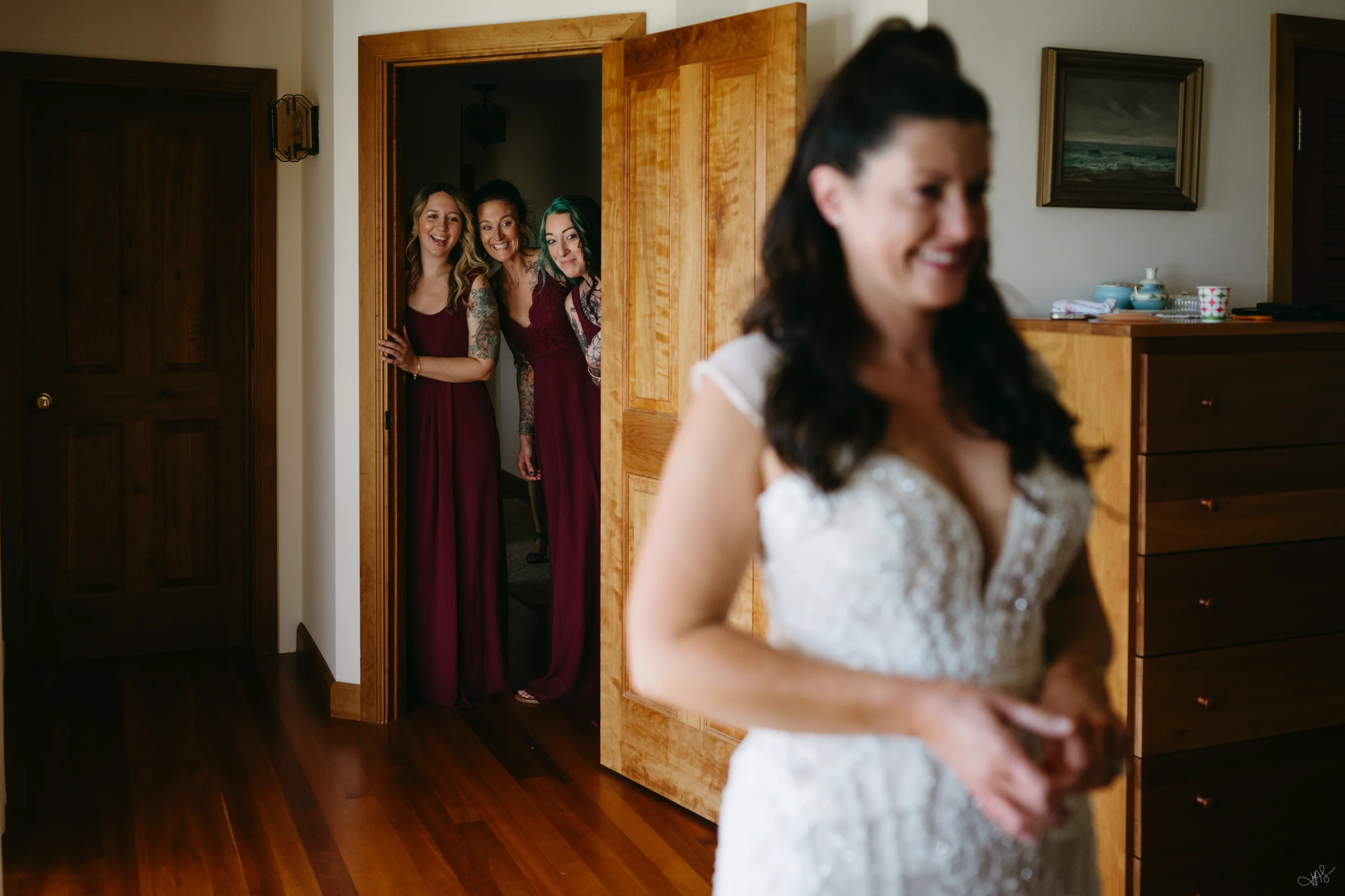 Four women peeking from behind a door, smiling at a woman in a white dress in a room with wooden furniture and a framed painting of an ocean scene.