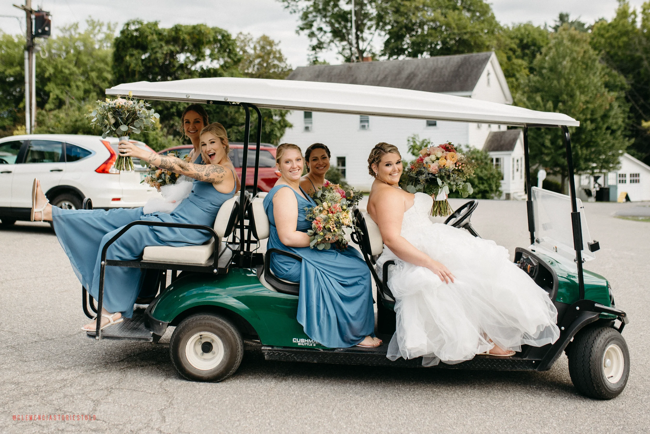 A bride in a wedding dress and five bridesmaids in blue dresses sitting on a golf cart, holding bouquets and smiling, with a background of parked cars and trees.
