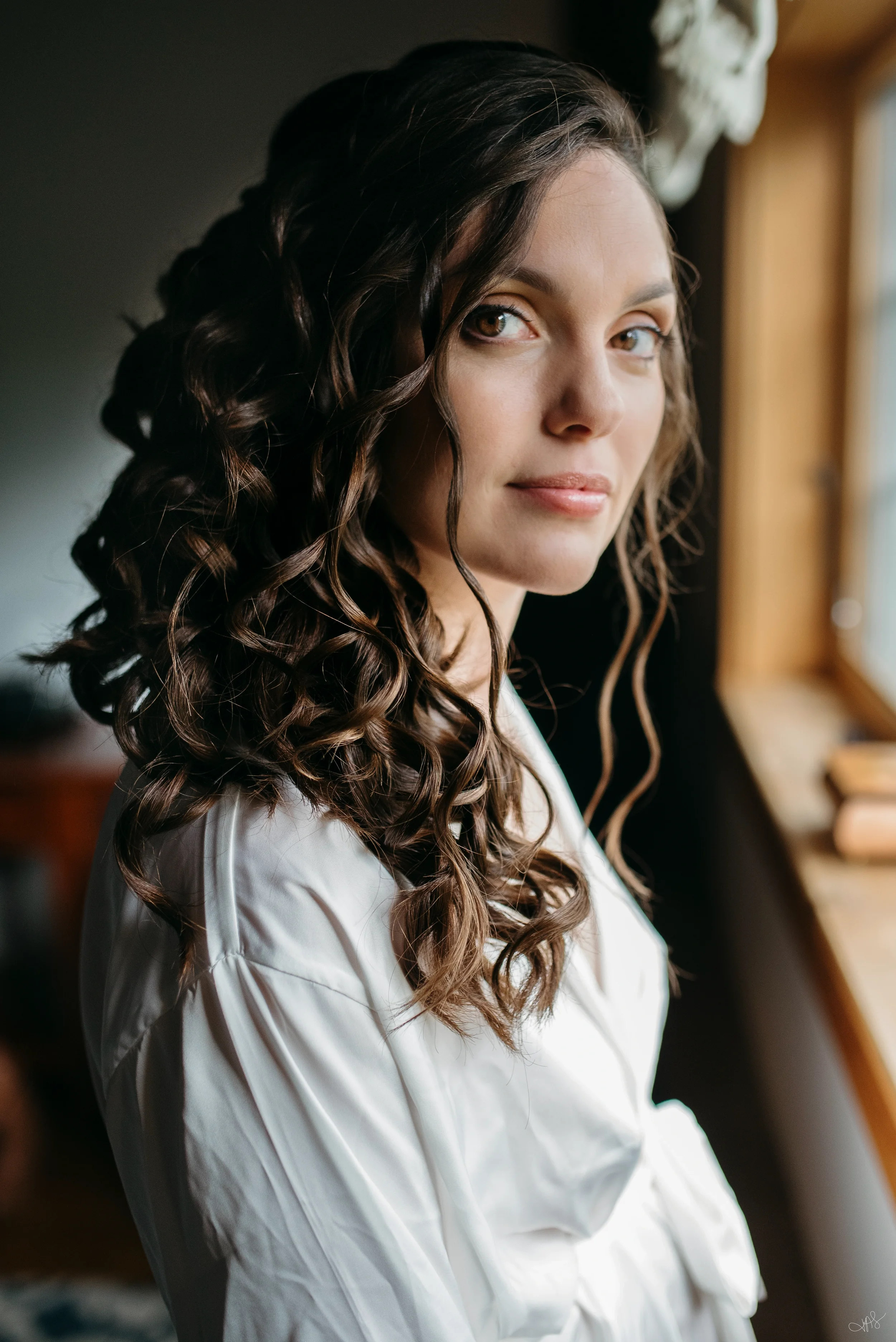 Close-up of a woman with long, dark, curly hair, wearing a white blouse, looking out a window with natural light on her face.