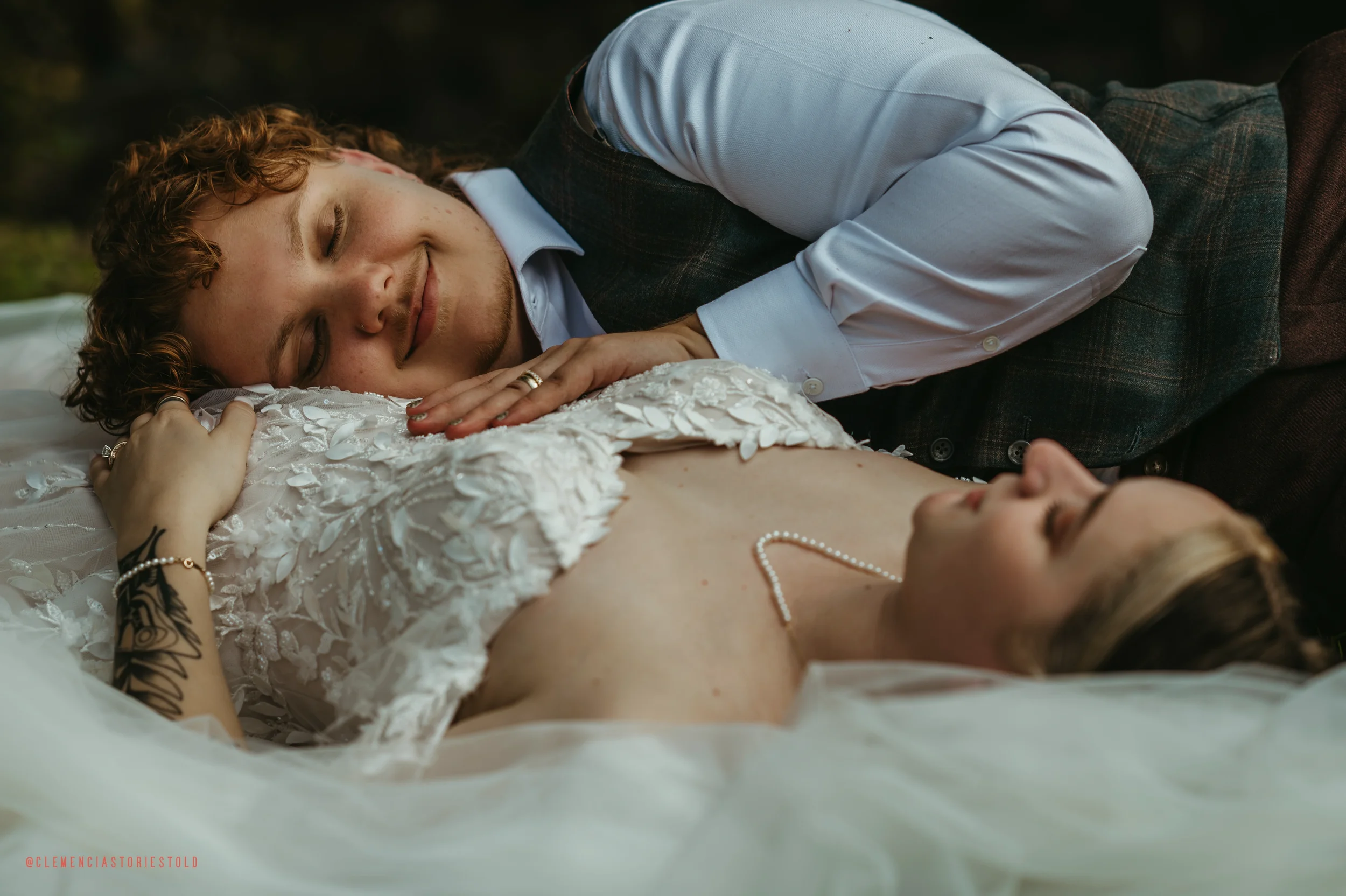 A couple lies on the ground, smiling and embracing. The woman is wearing a wedding dress and pearl necklace, and the man is dressed in a shirt and vest.