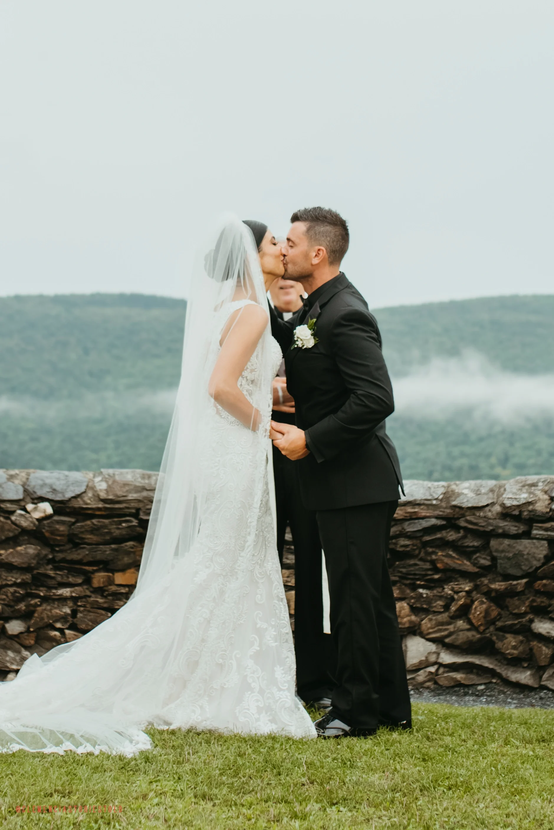 A bride and groom kiss during their outdoor wedding ceremony, with a scenic mountain view in the background and officiant standing behind them.