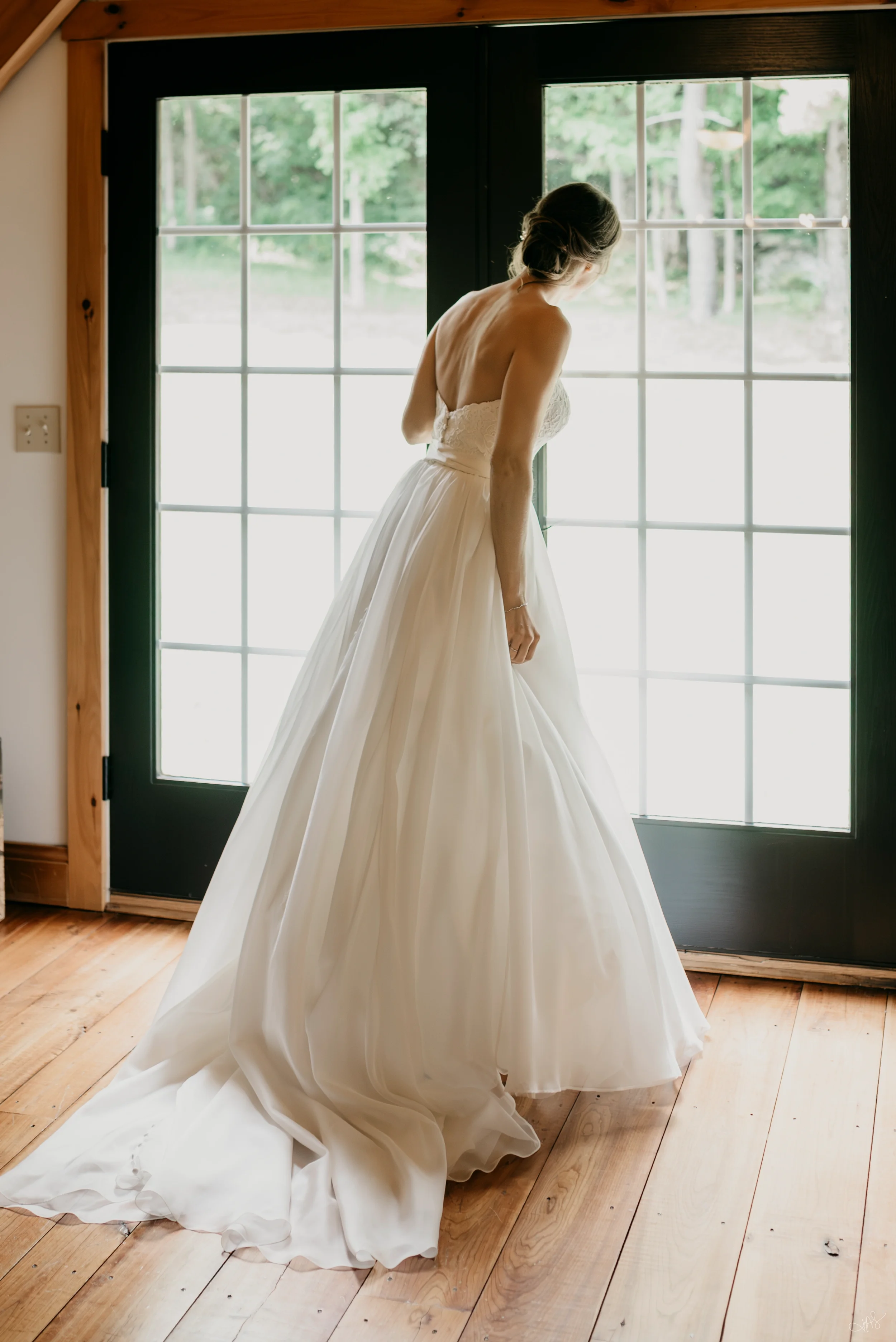 A woman in a wedding dress standing near a large window, looking outside. The dress is strapless with a fitted bodice and a full, flowing skirt that pools on the wooden floor. The woman has dark hair styled in an updo.