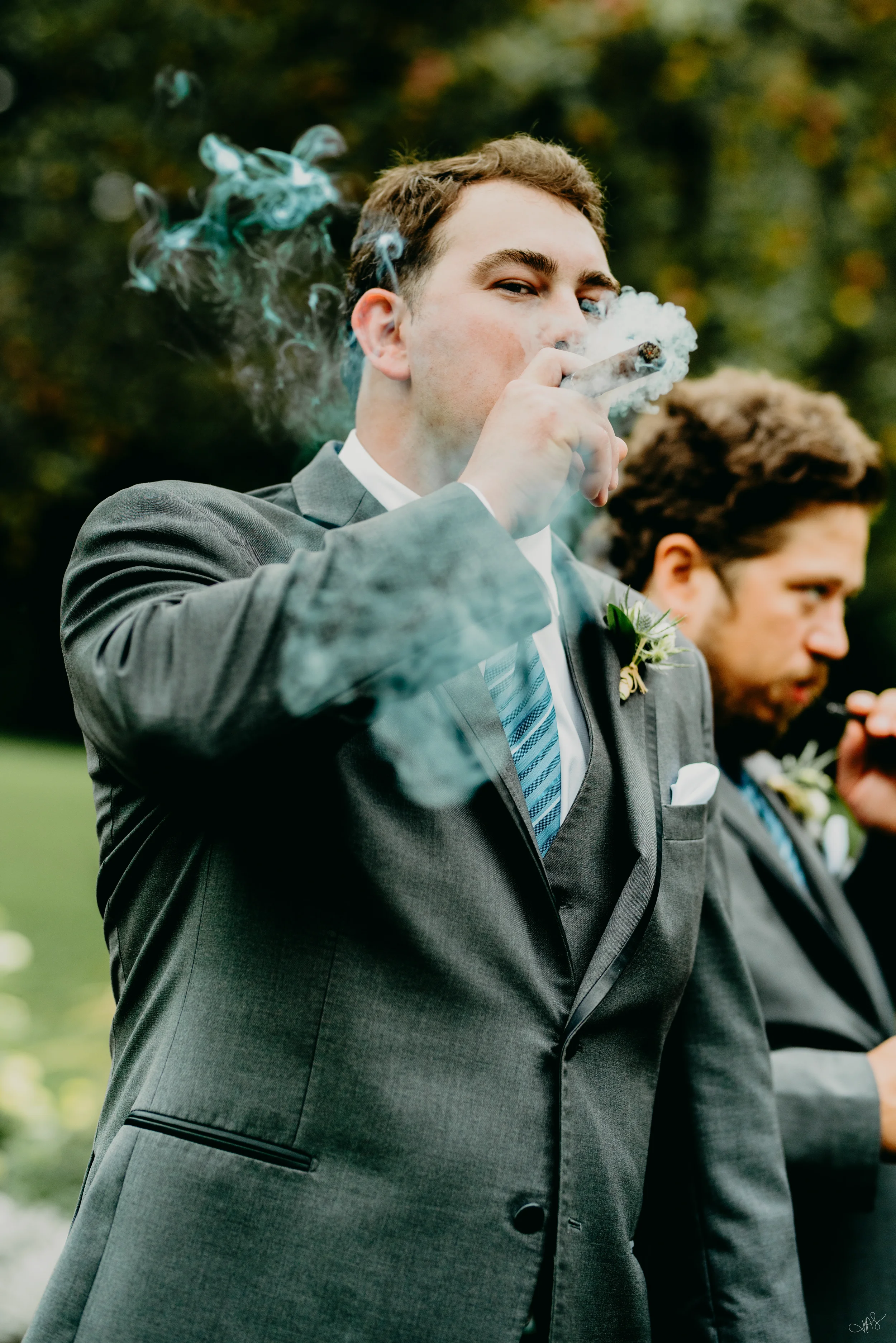 A man in a suit smoking a cigar outdoors during the daytime with blurred trees in the background.