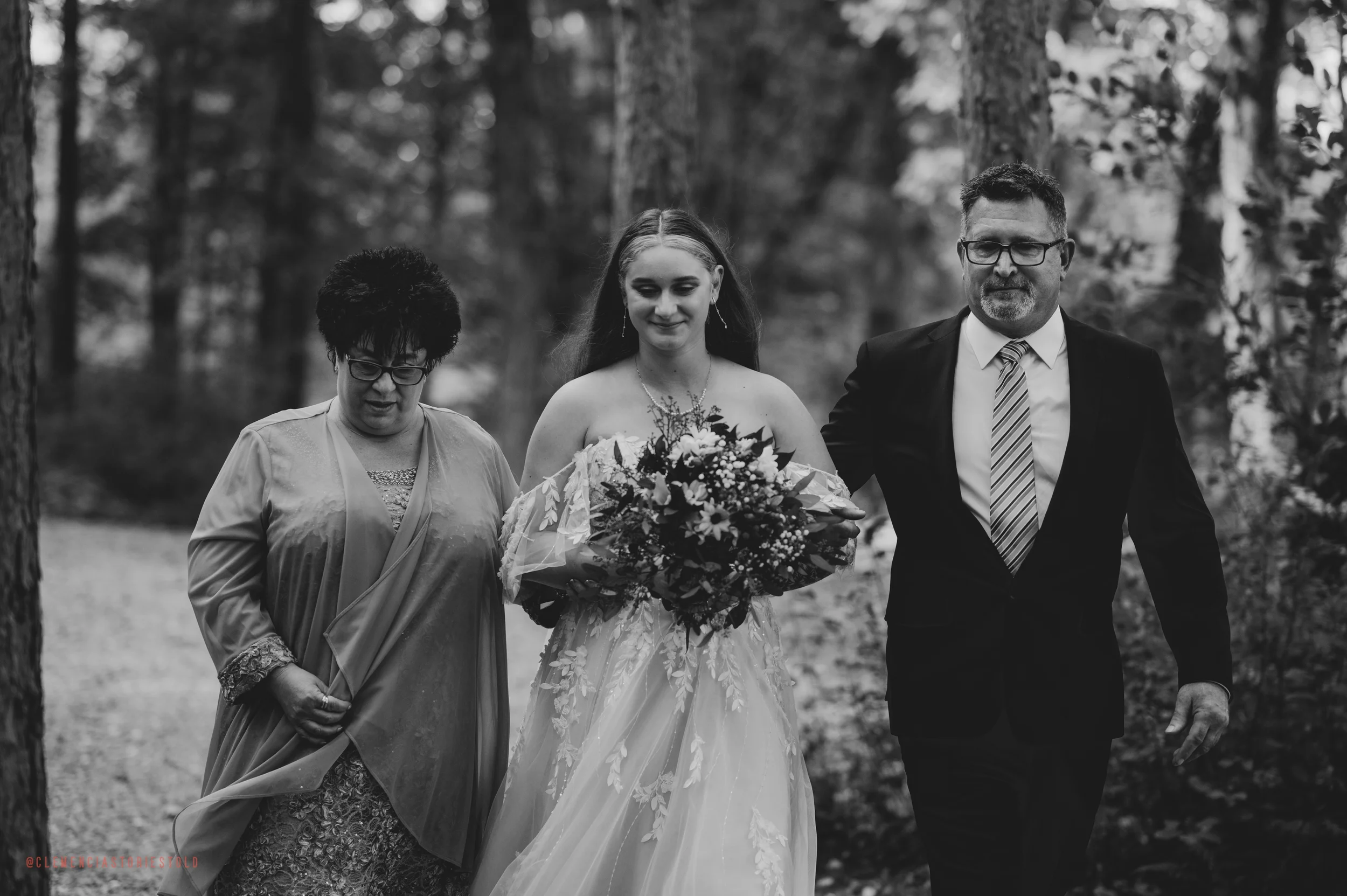 A black and white photograph of a bride walking outdoors with her parents in a wooded area. The bride is holding a large bouquet of flowers and smiling, with her parents on either side of her.