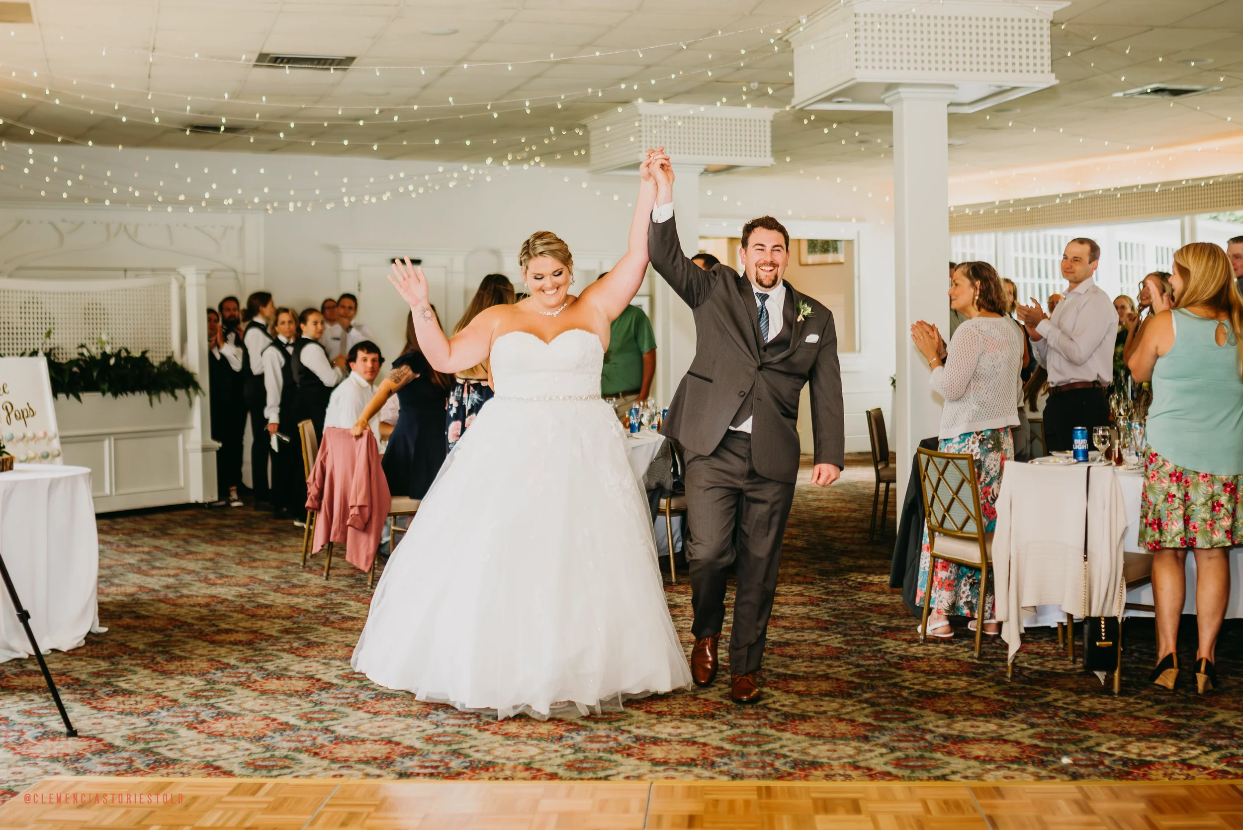 Bride and groom holding hands and celebrating their wedding dance in reception hall with guests clapping and smiling.