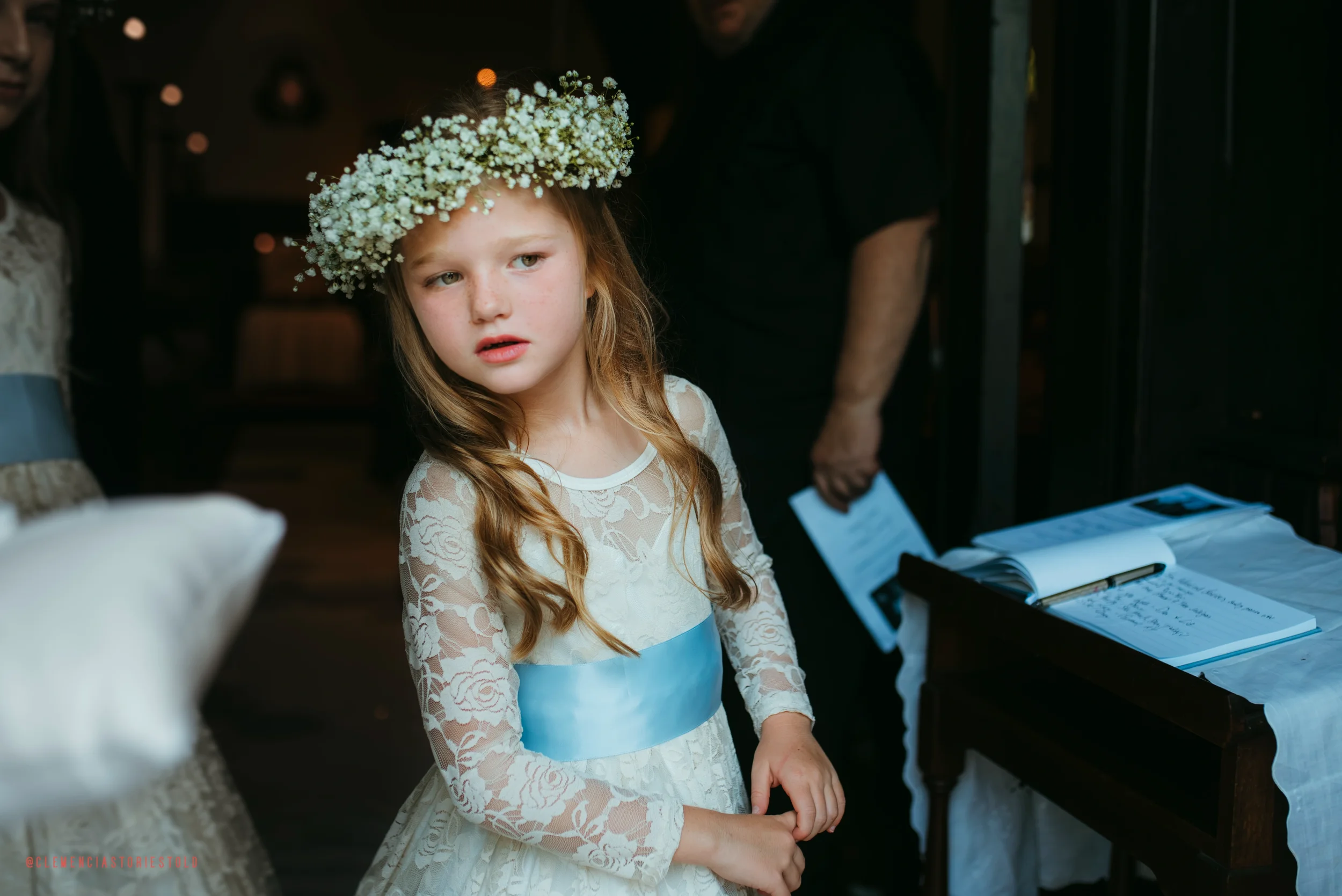 A young girl with long wavy hair wearing a white lace dress with a light blue sash and a crown of small white flowers on her head, looking thoughtfully at something outside the frame.
