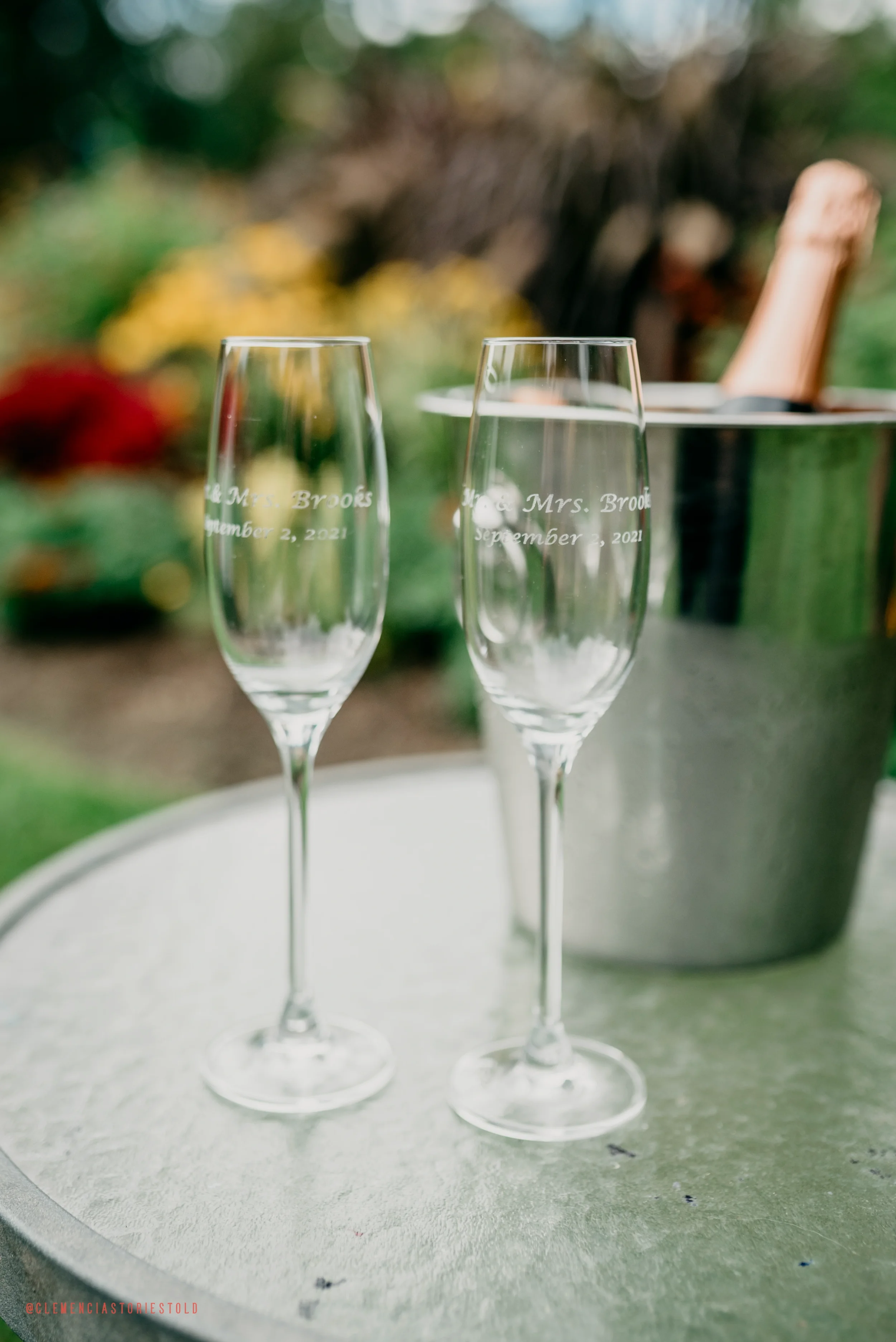Two empty champagne glasses with engraved text reading 'Mr. & Mrs. Brooks September 2, 2021,' placed on a round metallic table outdoors. A bucket with a bottle of champagne is in the background, with blurred green foliage.