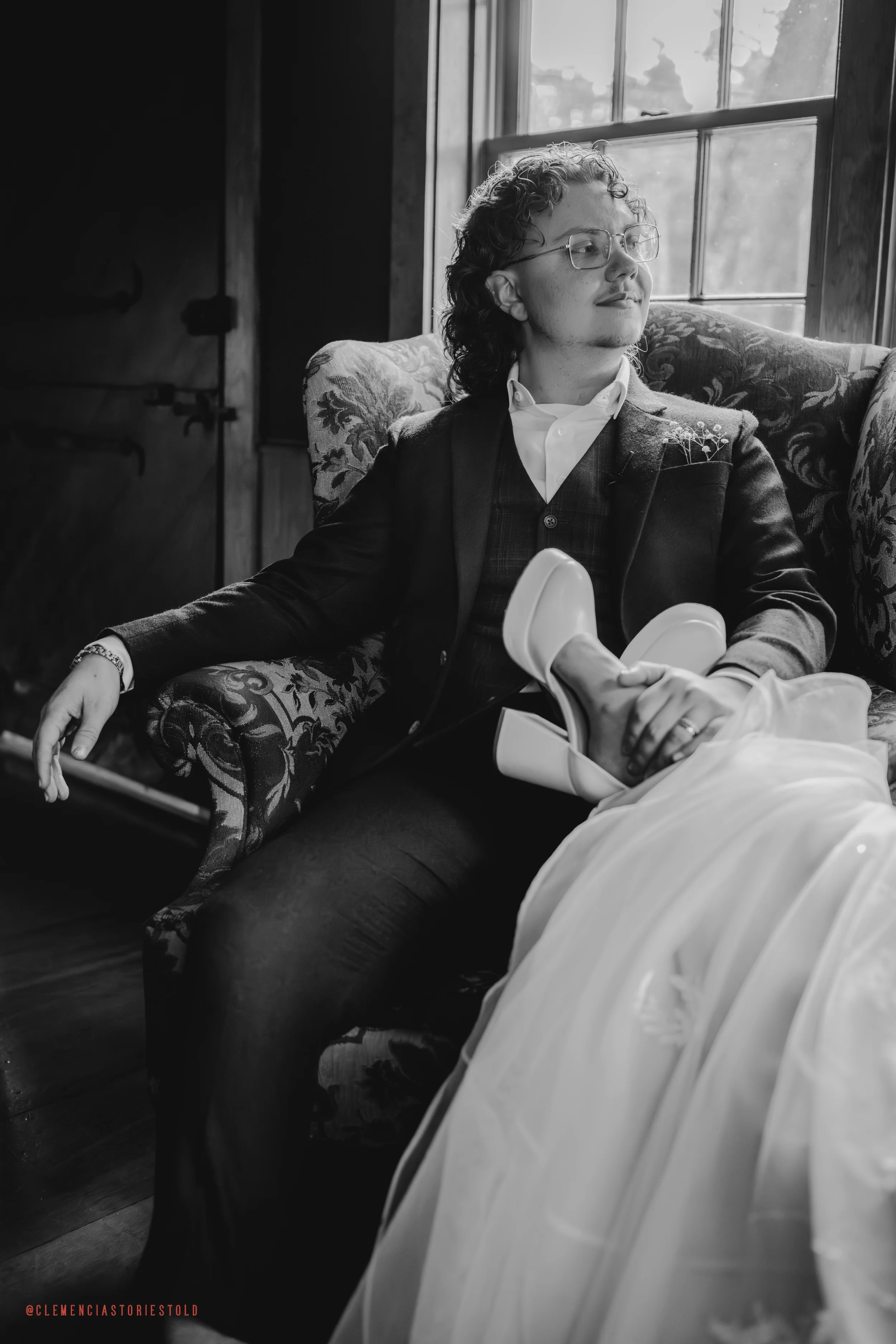A person with curly hair and glasses sitting in a vintage-looking armchair, wearing a suit. They are gazing thoughtfully out a window with natural light coming in, and their hands are resting on their lap, one hand on a white dress or gown.