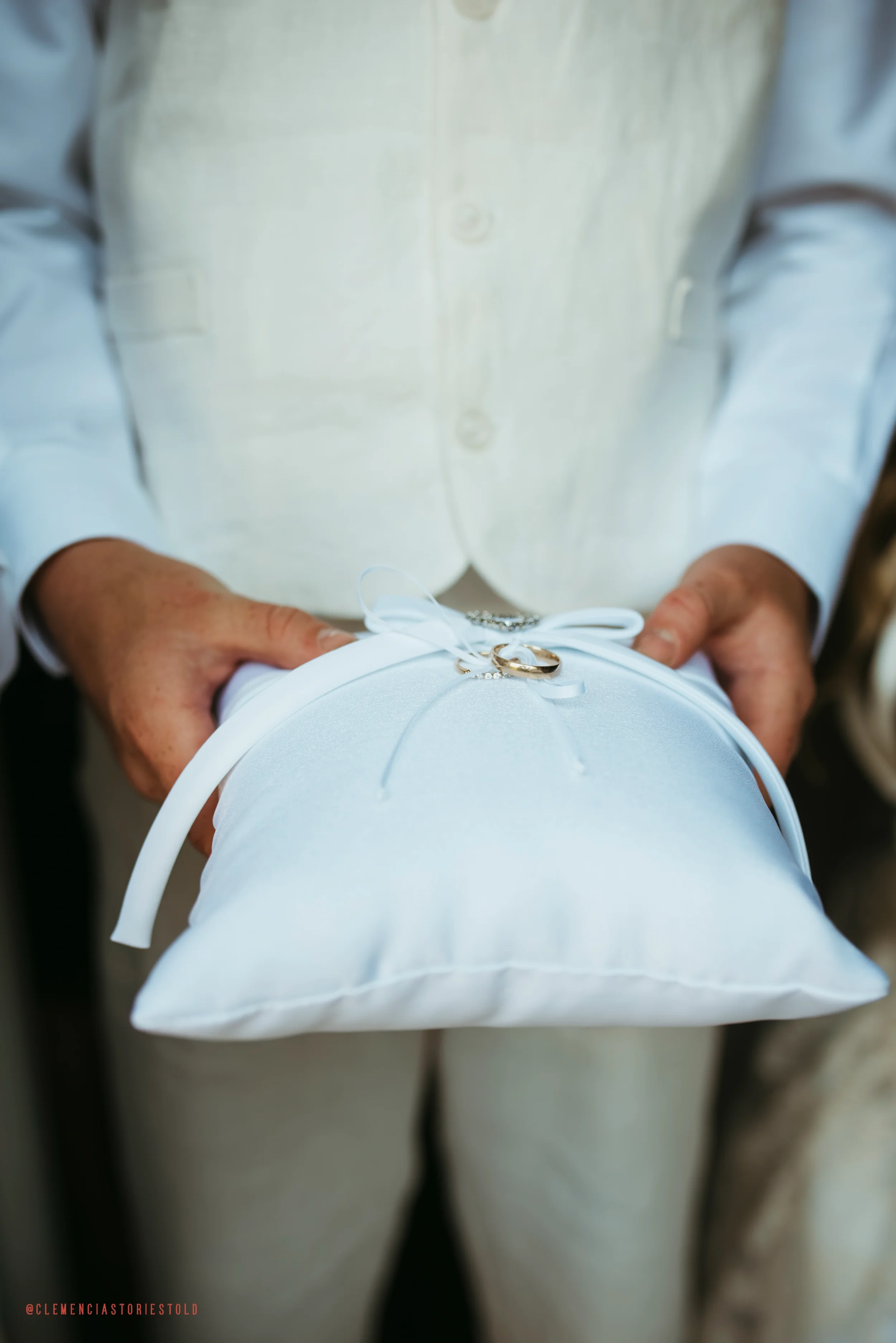 Person holding a white pillow with wedding rings on top, dressed in light-colored clothing.