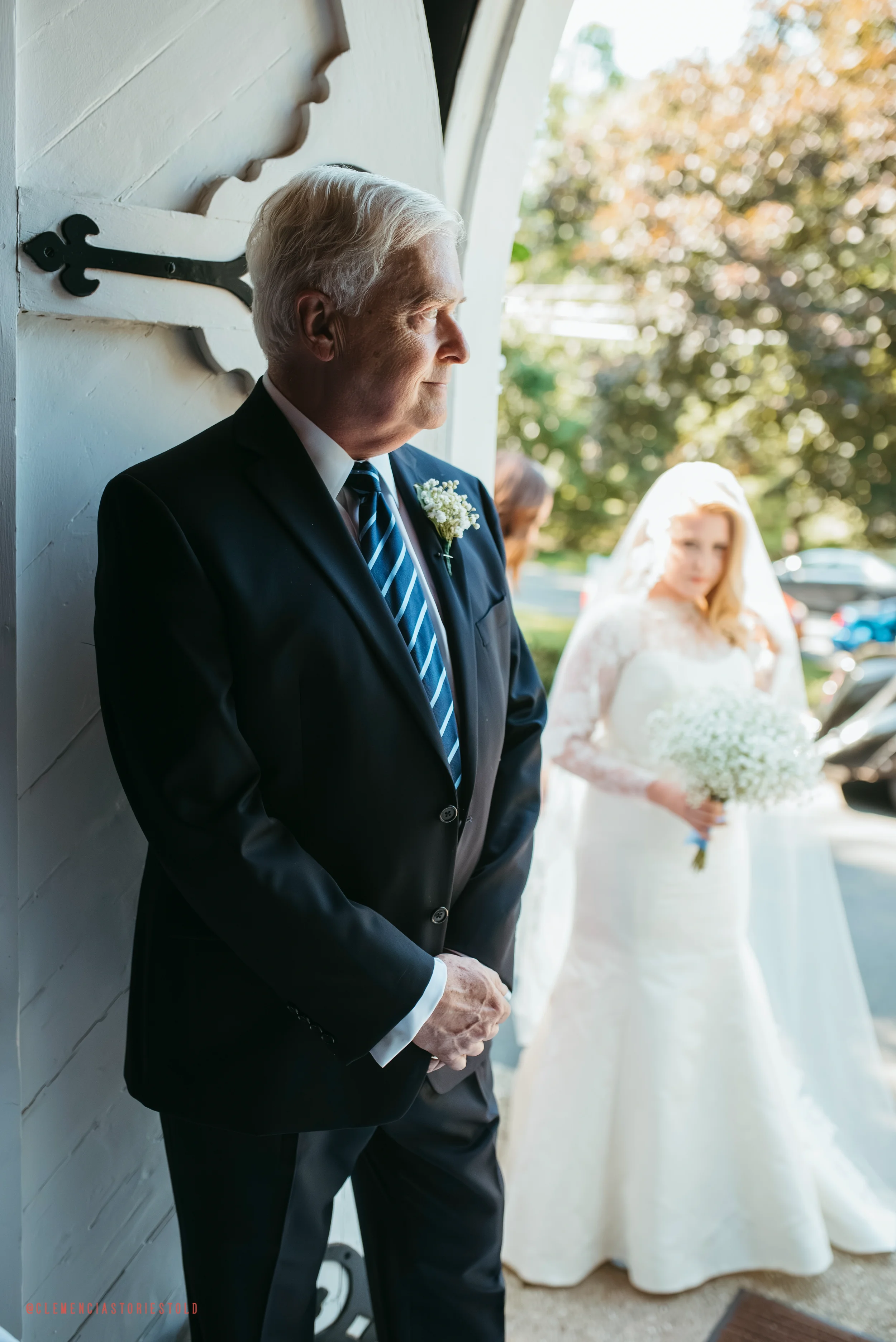 An elderly man in a black suit with a blue striped tie, standing with hands clasped, looking out a window at a bride in a white wedding gown holding a bouquet of white flowers.