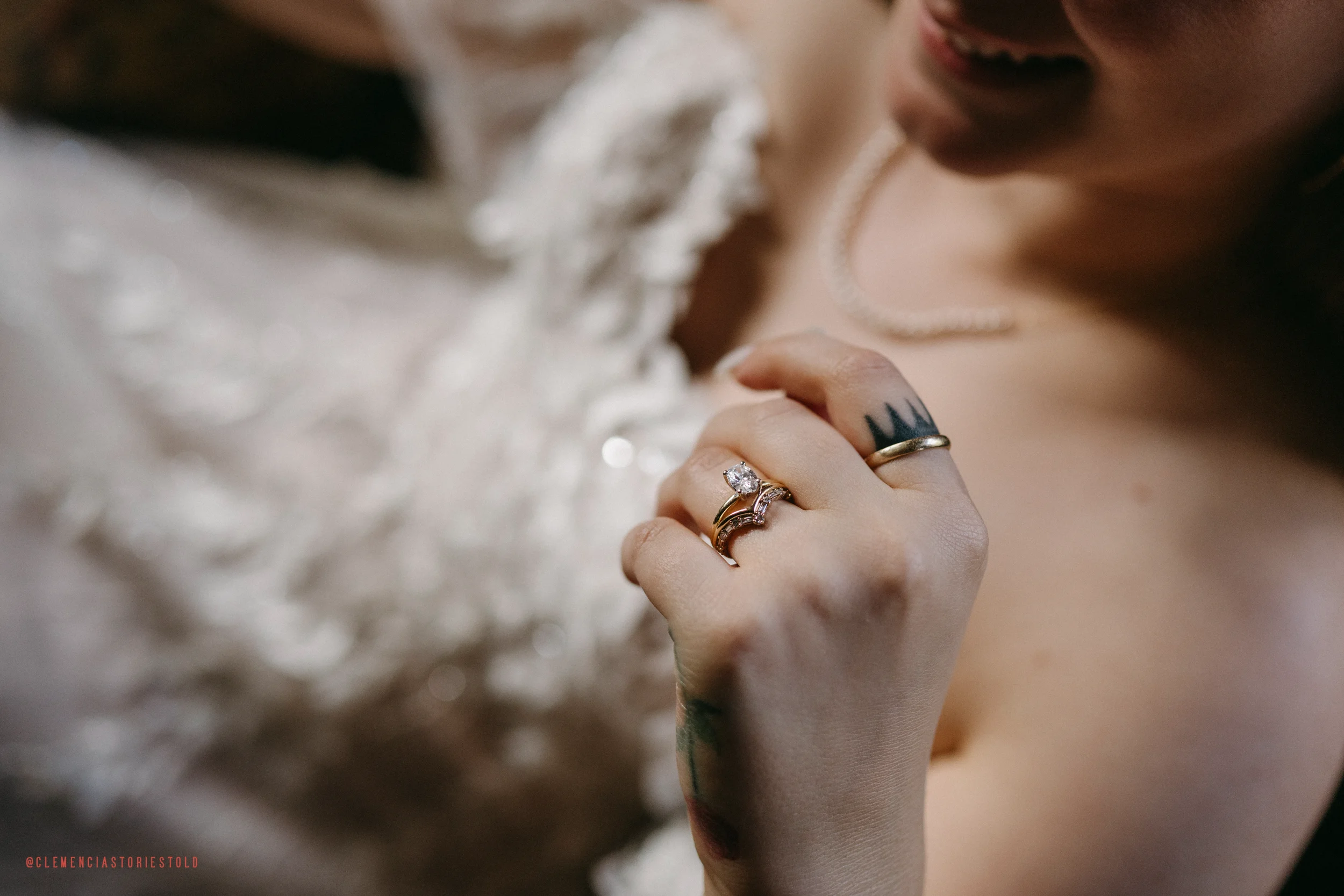 Close-up of a person with tattoos on their hand, wearing a wedding ring and a prominent diamond engagement ring, their lips slightly parted, dressed in a white lace garment, holding their chin with fingers.