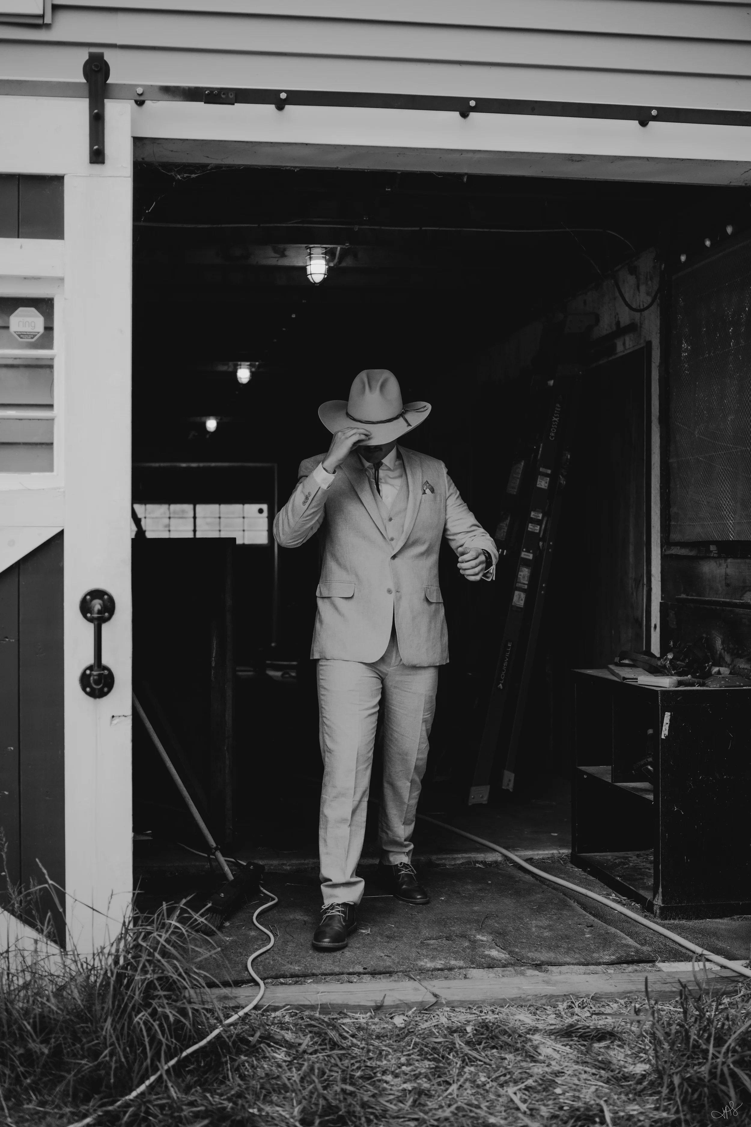 A man in a suit and wool hat stands at the entrance of a garage or workshop, adjusting his hat with his hand. The setting appears industrial or artistic, with visible power cords, tools, and a partially open garage door.