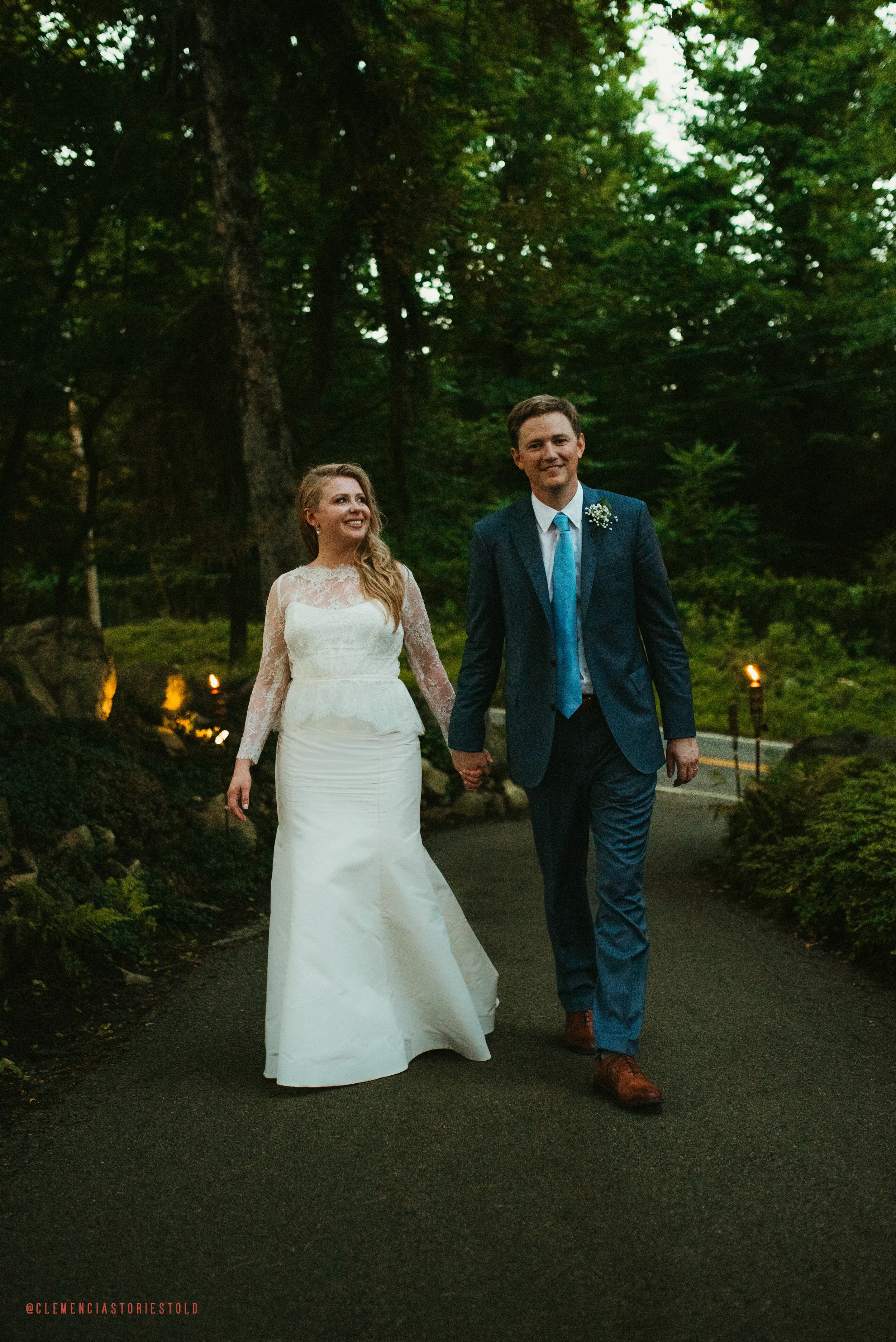 A bride and groom walking hand-in-hand on a wooded path during their wedding, smiling and looking happy.