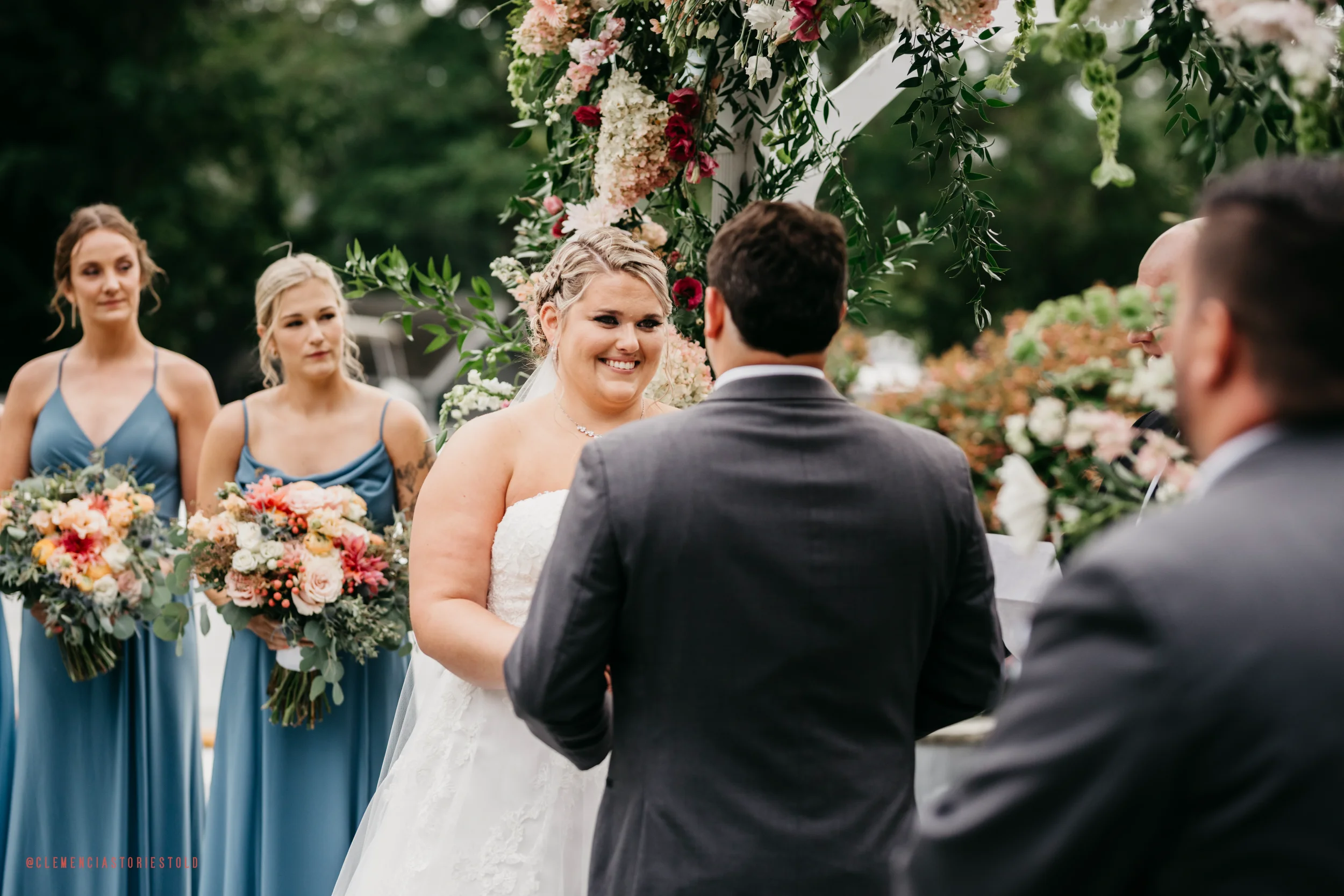 Bride and groom exchange vows outdoors during wedding ceremony, with bridesmaids holding bouquets in background.