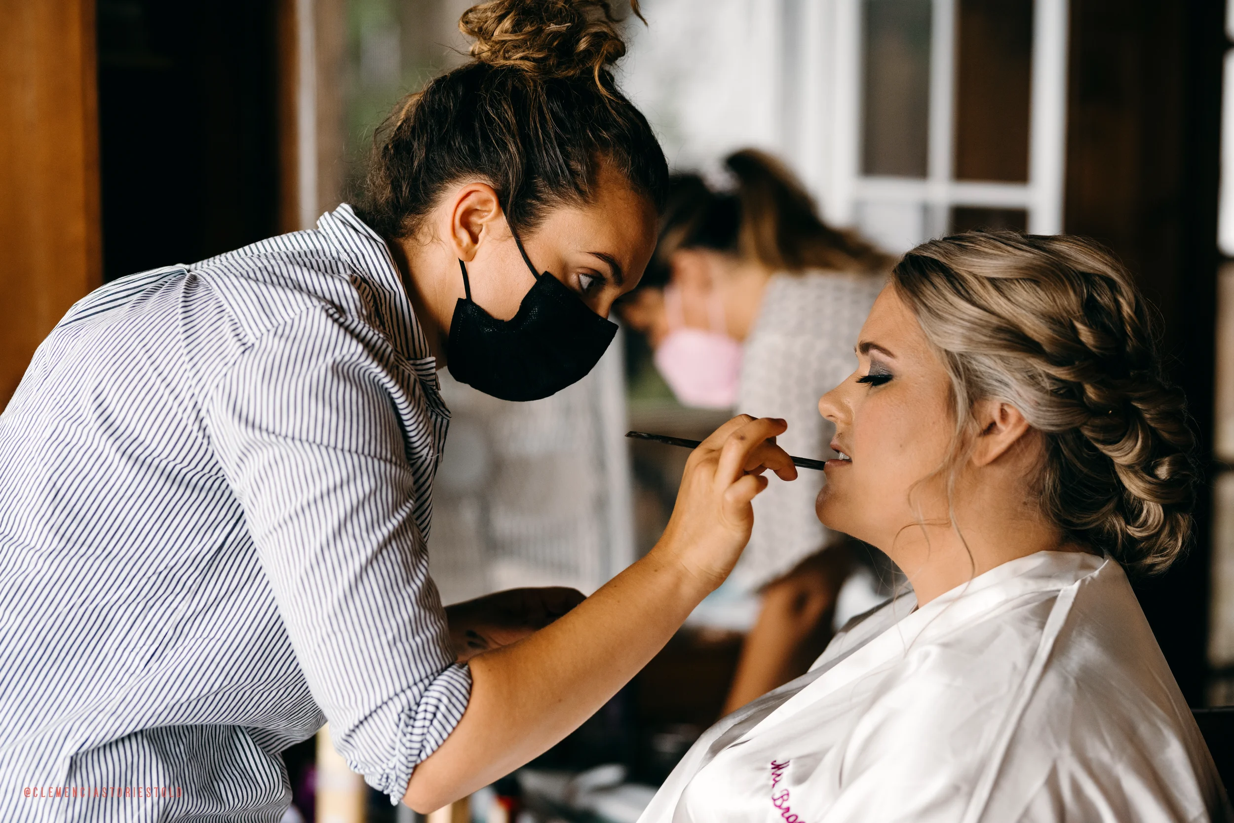 Makeup artist in black face mask applying lipstick to a woman with blonde hair in a salon.