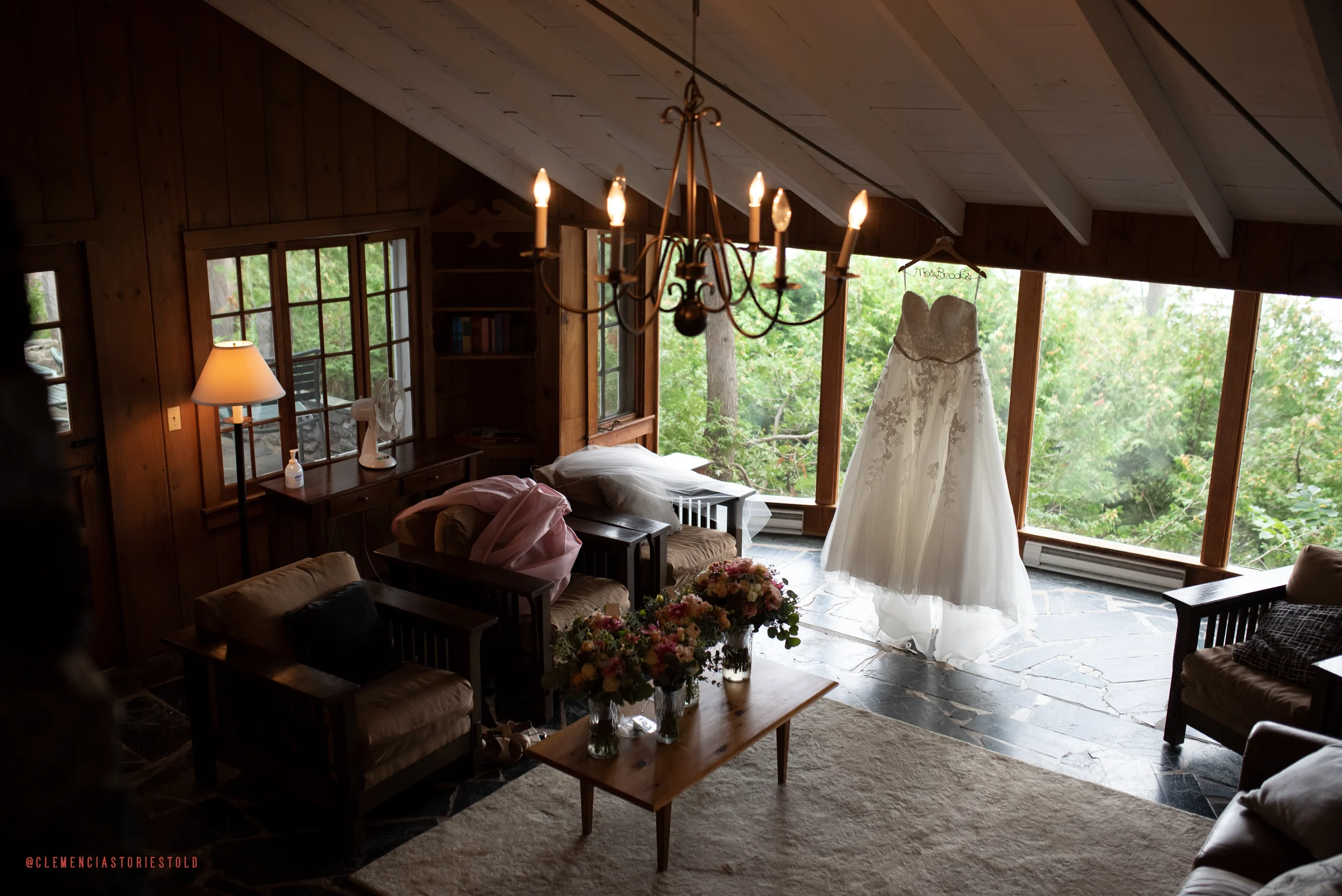 A wedding dress hanging in a room with large windows viewing a green forest outside. The room has wood-paneled walls, a chandelier, a sofa, armchairs, a table with flower arrangements, and a lamp.