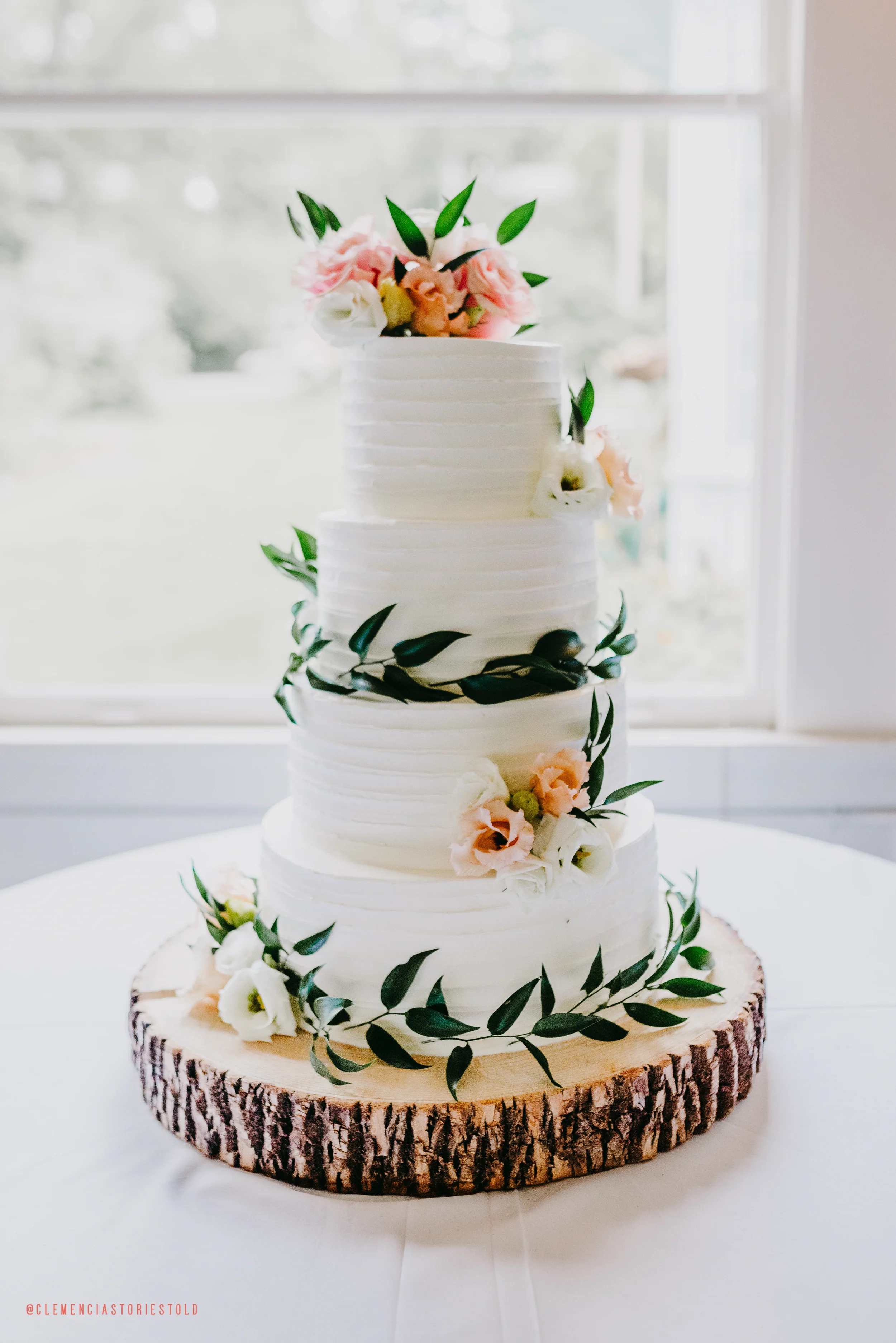 A four-tiered white wedding cake decorated with flowers and green leaves, placed on a wooden slice base, near a window with natural light.