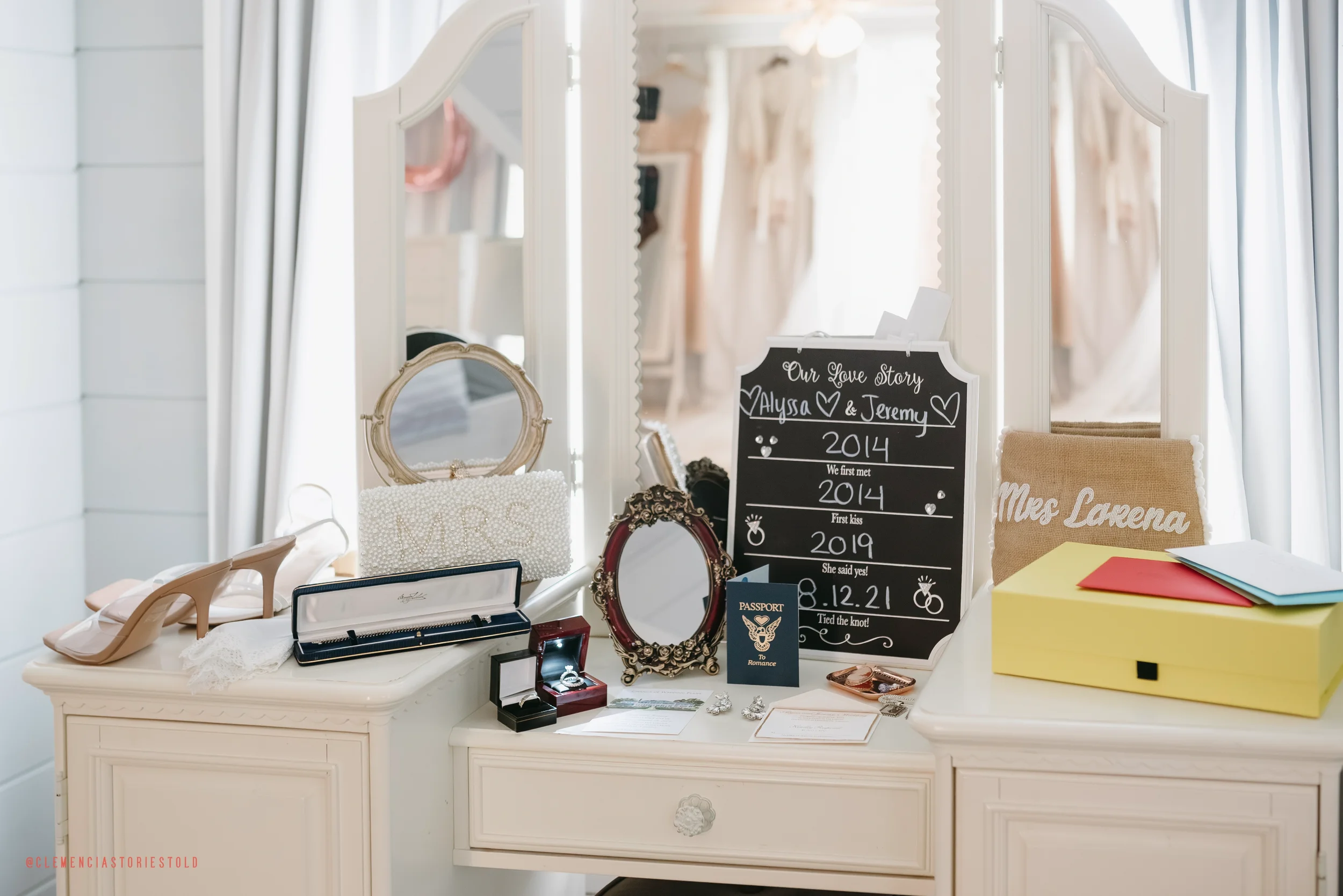 Wedding memorabilia on a white dresser, including a pair of nude high heels, jewelry boxes, vintage mirrors, a chalkboard with wedding details, and a beige pillow with white text, with a large mirror behind reflecting hanging wedding dresses.