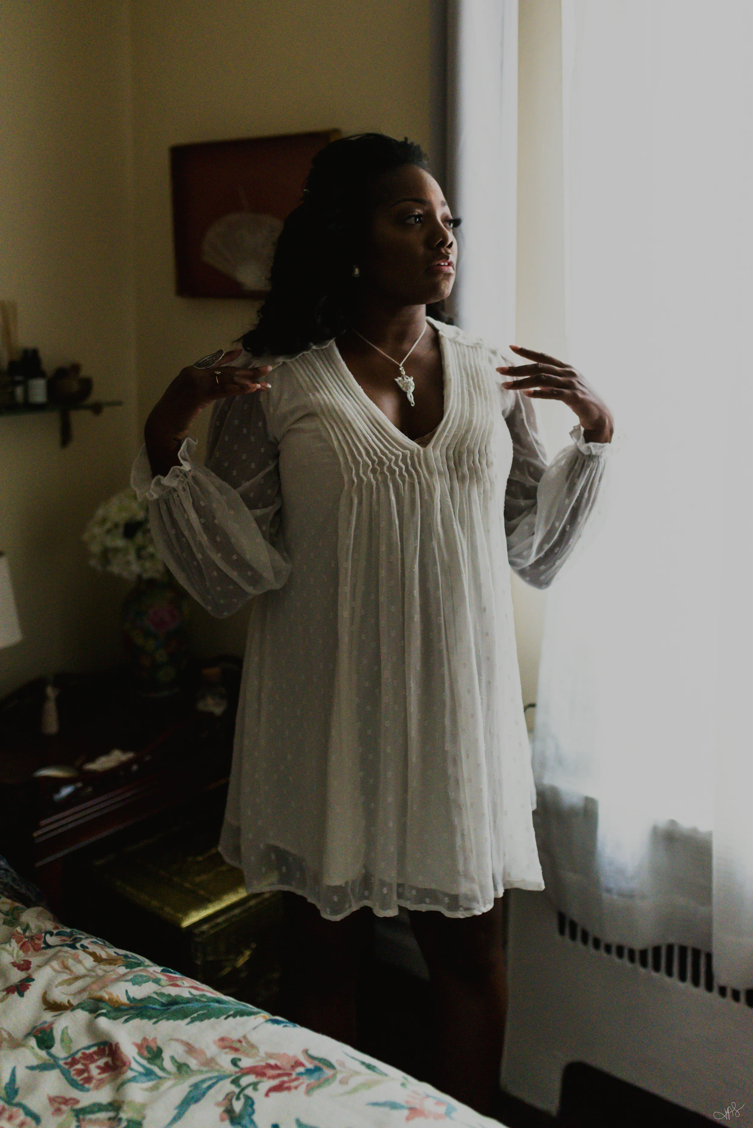 A woman in a white dress with intricate embroidery stands by a window, looking outside with her hands near her collarbone. The room has a dark wooden nightstand with bottles and framed photos, a floral bedspread, and light-colored curtains.