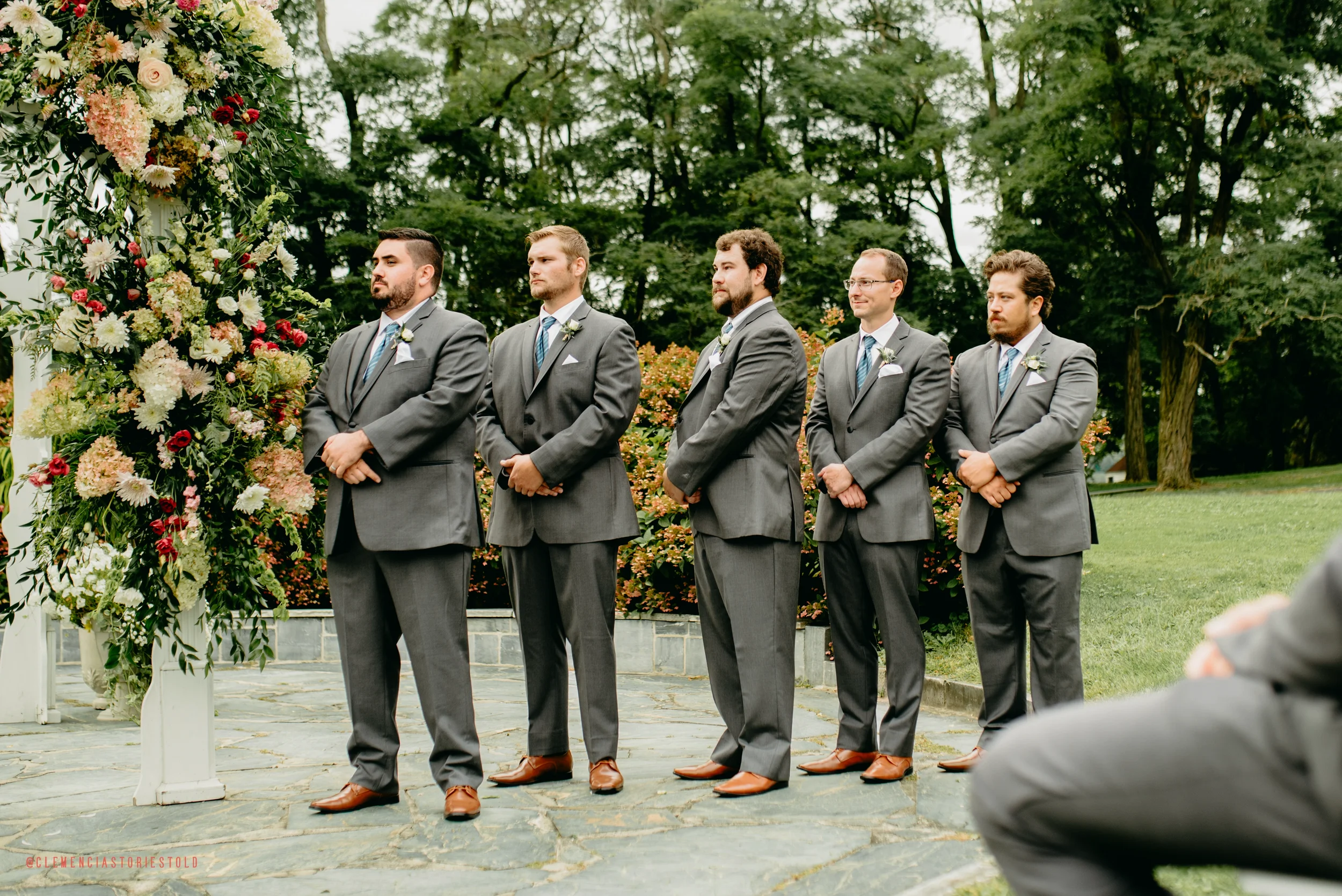 Groom and five groomsmen standing outdoors during a wedding ceremony, dressed in gray suits with ties, in front of a floral arch and surrounded by trees and greenery.