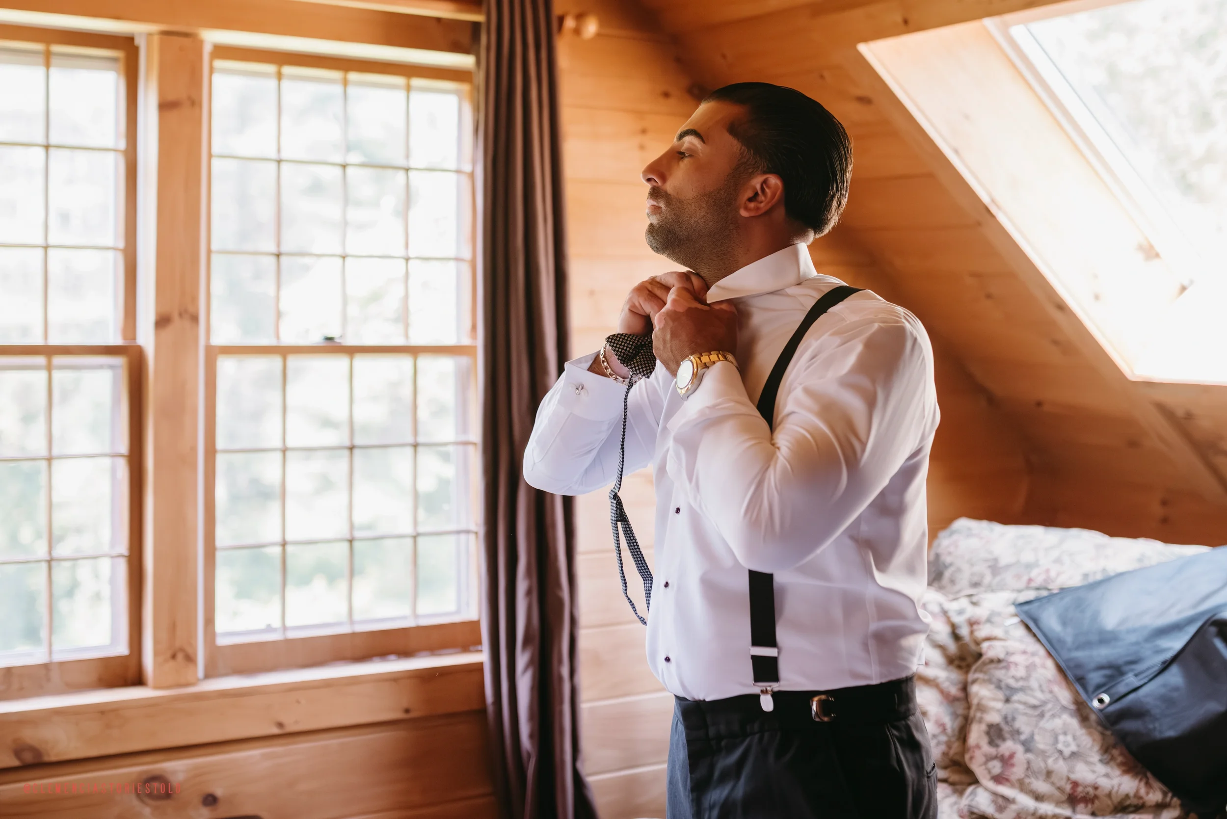 A man adjusting his shirt collar in a wooden room with large windows and a skylight, dressed in formal attire with suspenders and wearing a watch.
