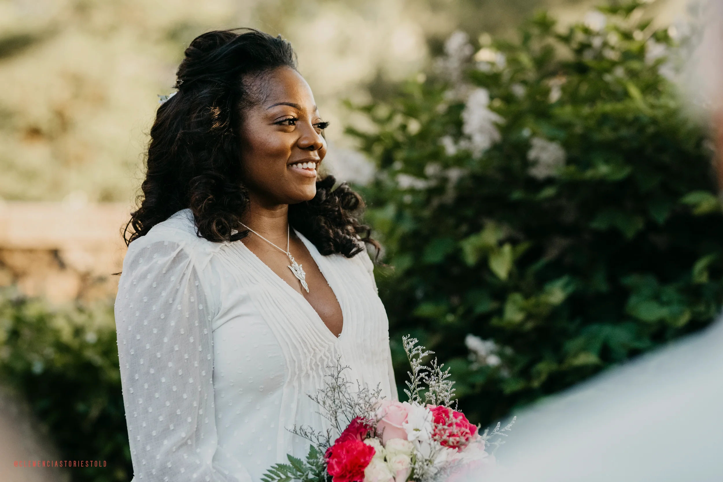 A woman with dark curly hair smiling, holding a bouquet of pink and white flowers, standing outdoors with greenery in the background.