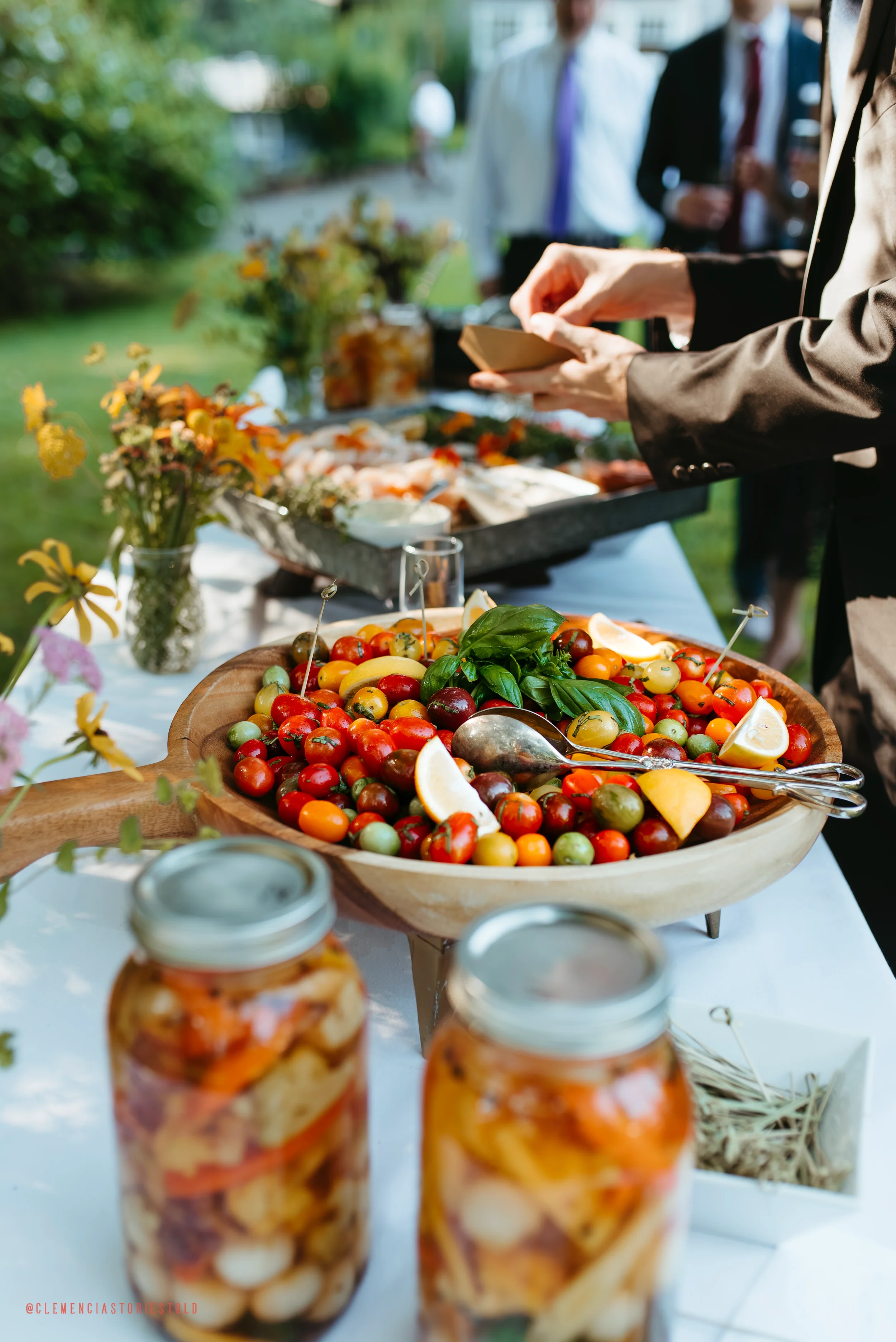 A table with a large wooden bowl of assorted cherry tomatoes, lemon wedges, and basil, alongside jars of pickled vegetables at an outdoor gathering with people in the background.