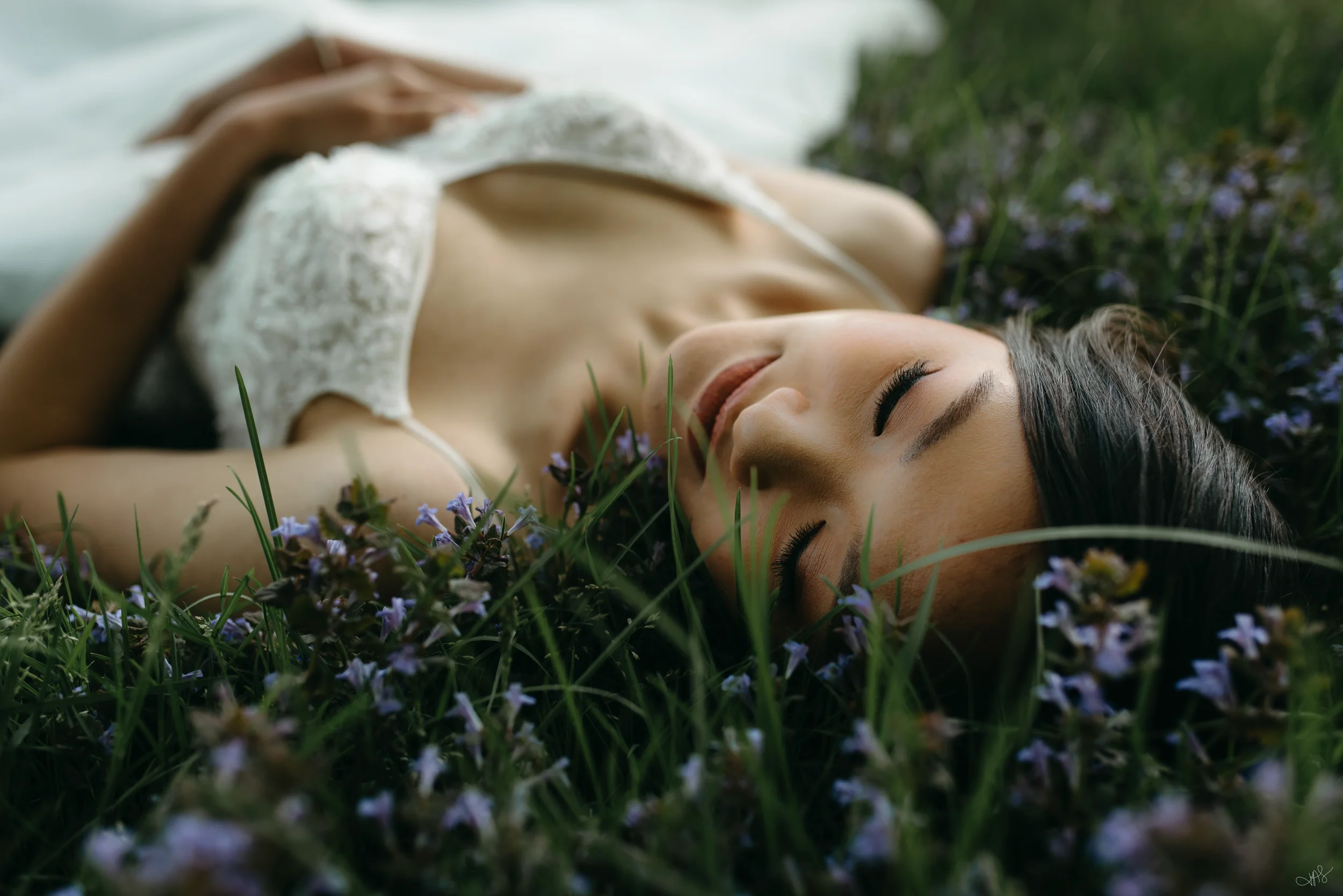 A young woman lying on her back in a field of small purple flowers with her eyes closed, wearing a white lace dress.
