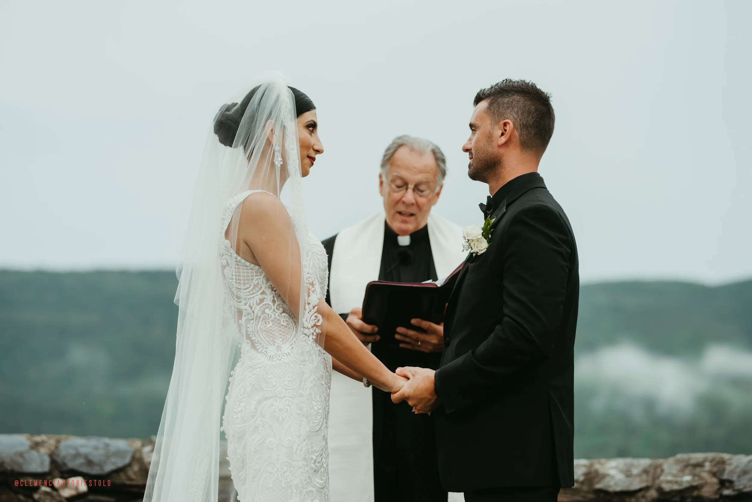 A bride and groom holding hands and exchanging vows during a wedding ceremony outdoors, with a officiant officiating, in a scenic natural setting with mountains in the background.