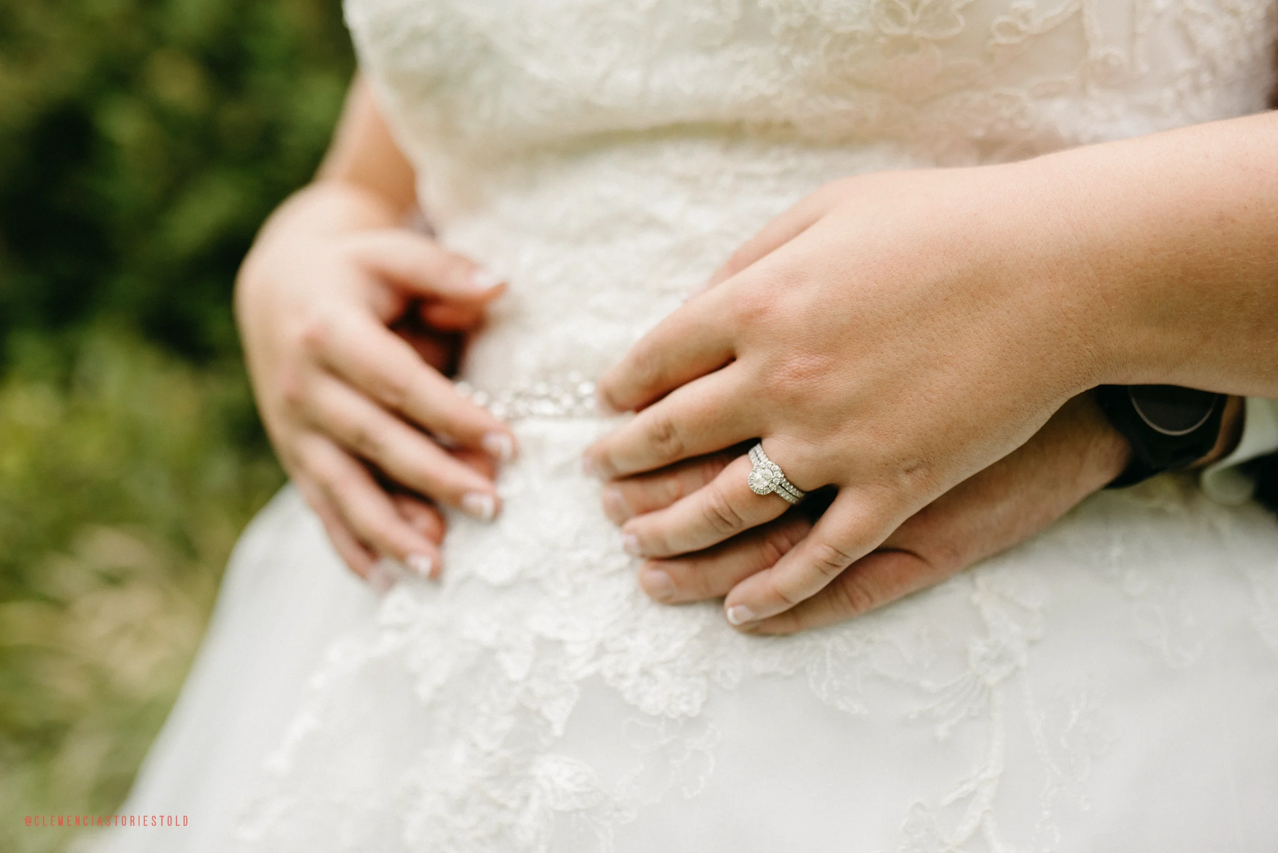 Close-up of a bride's and groom's hands, with the bride wearing an engagement ring and wedding band, gently resting on her wedding dress, with a blurred outdoor background.