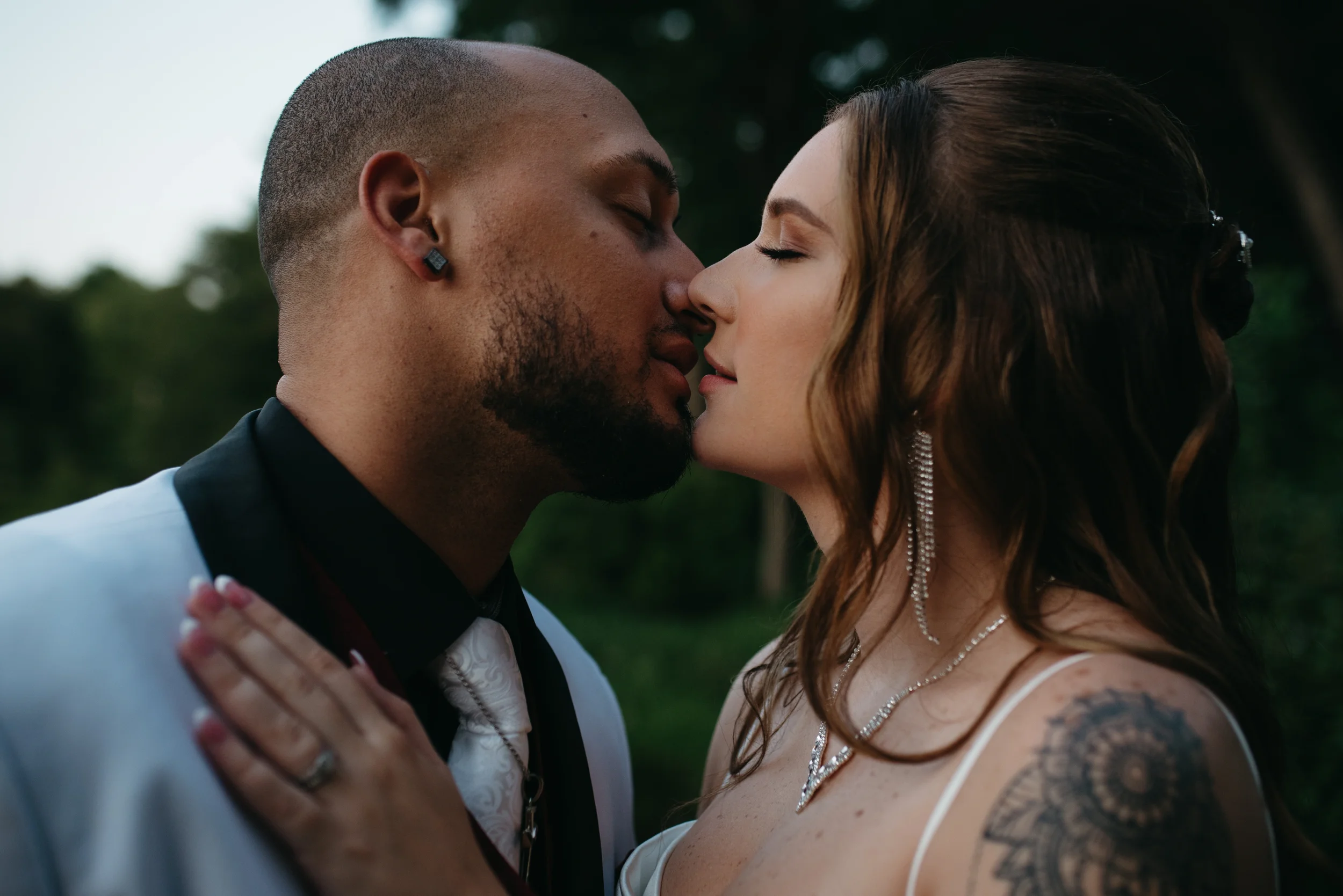 A couple, a man and a woman, about to kiss outdoors surrounded by trees. The man has a shaved head, beard, and is wearing a tuxedo with an earring. The woman has long brown hair, closed eyes, and tattoos on her shoulder and arm, wearing a white dress with jewelry.