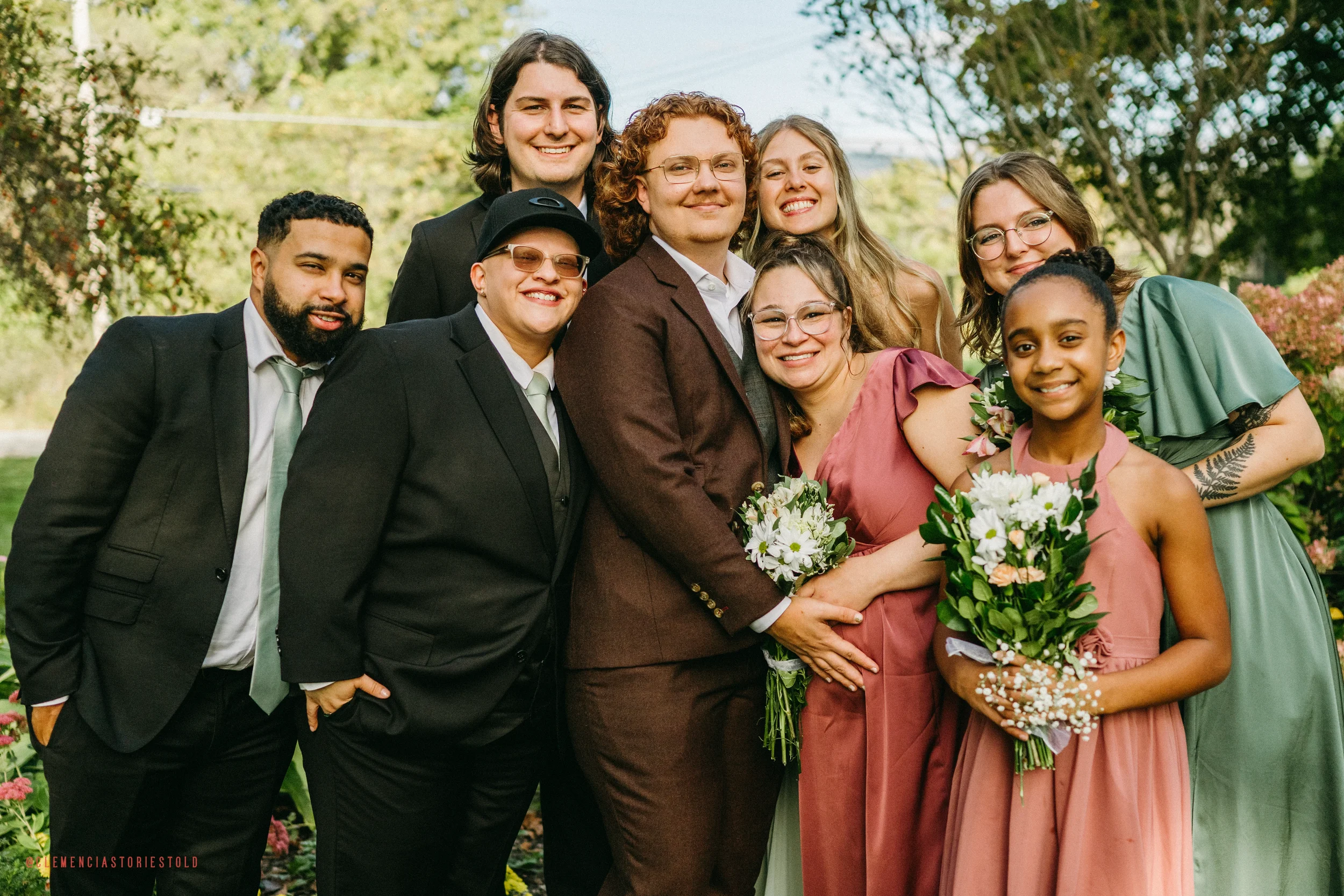 A group of nine people, including adults and children, standing together outdoors during a celebration, smiling and holding flowers, with trees and greenery in the background.