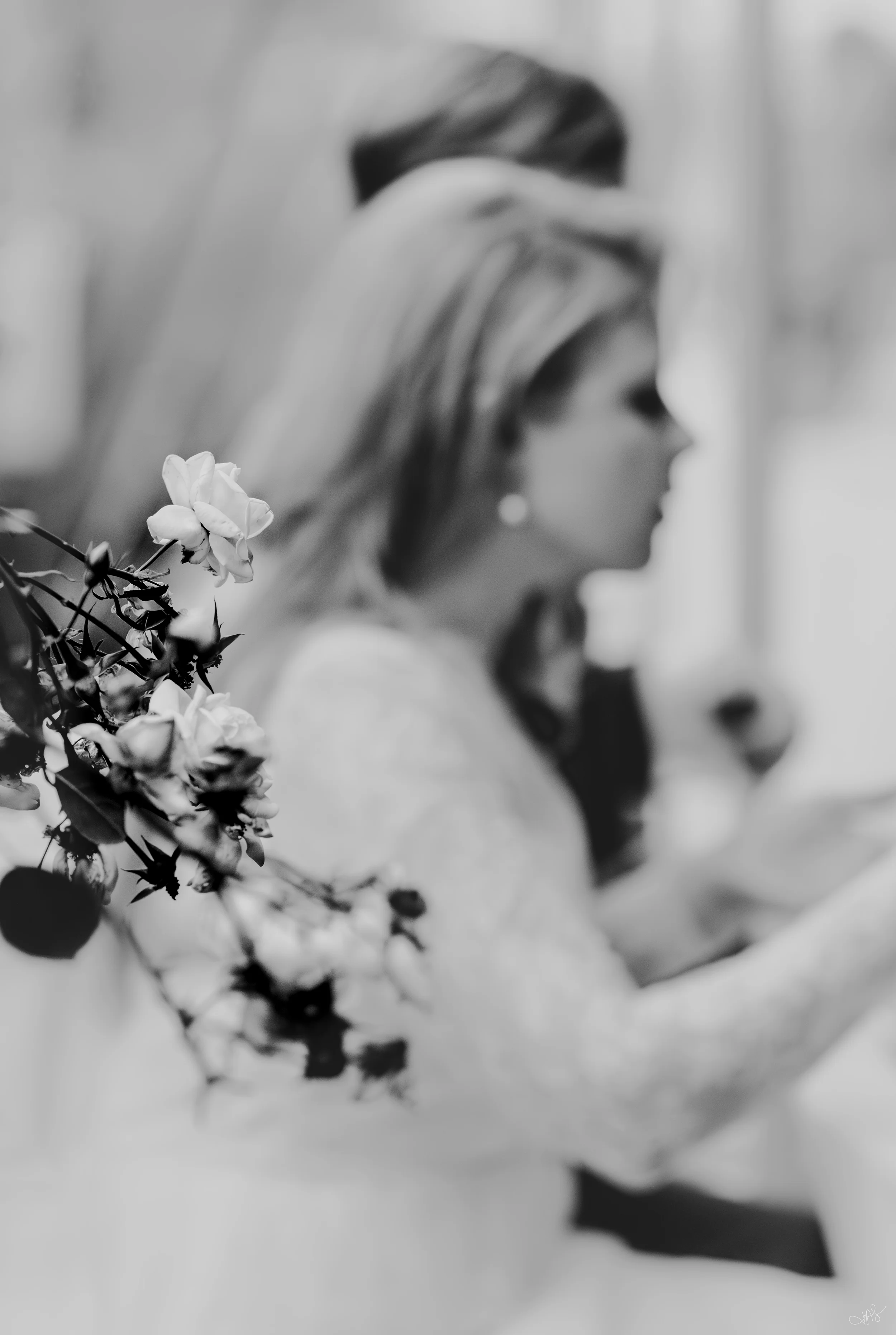 A woman with long hair touching her face, standing near a branch of white flowers in a blurred background, black and white photograph.